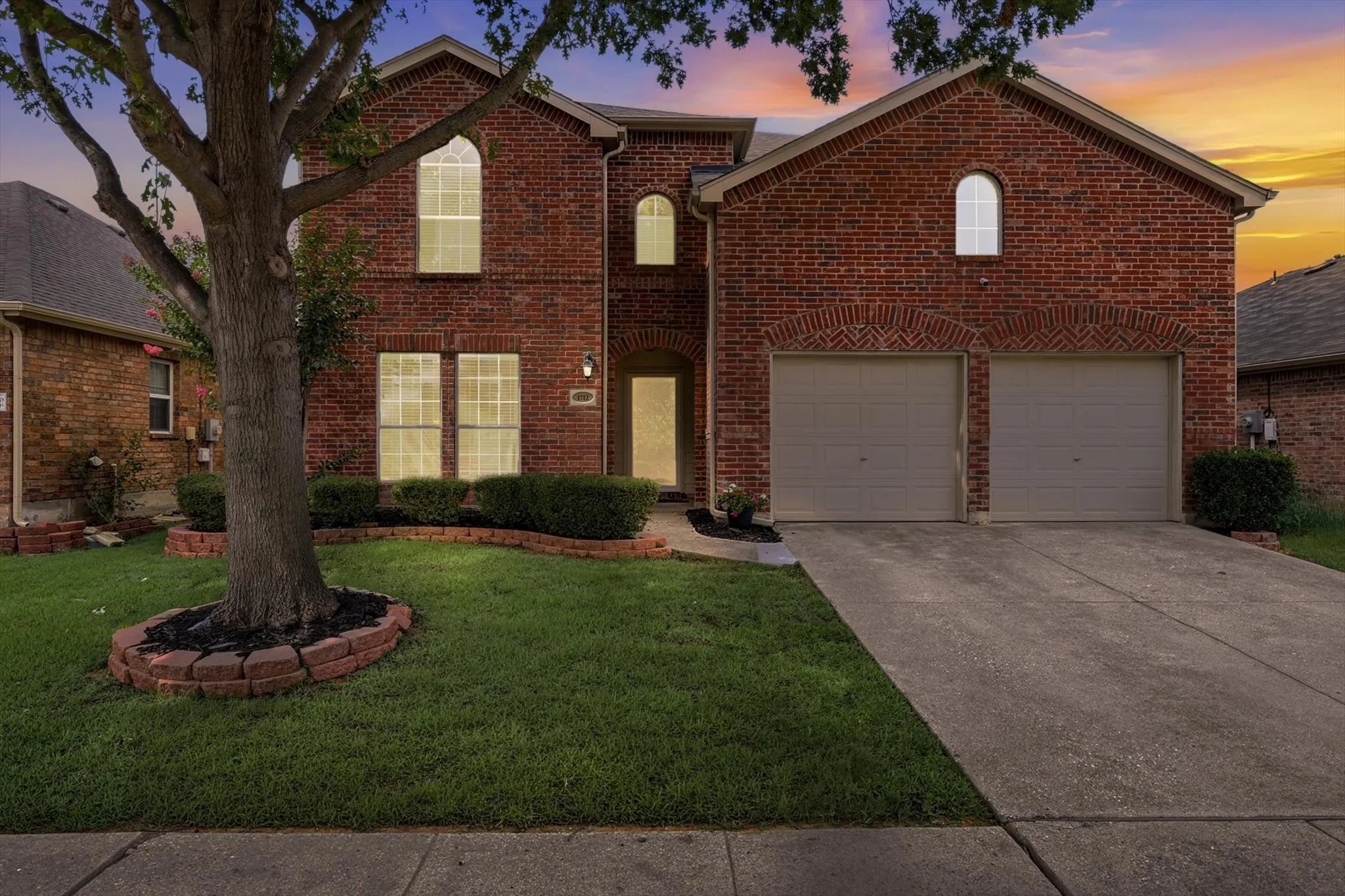 Evening-Brick exterior residence featuring a two-car garage with arched brick detailing, a concrete driveway, and a front lawn with a mature tree