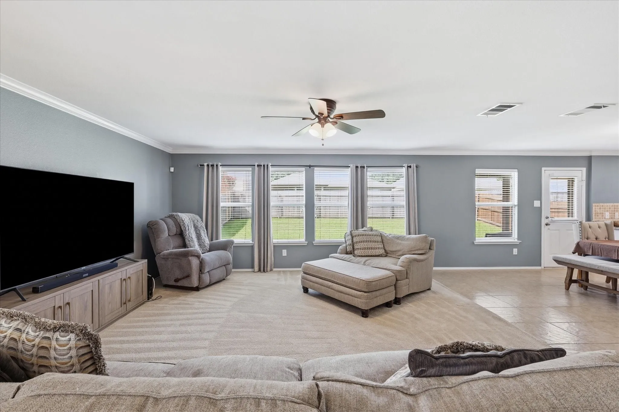 Spacious living area featuring carpet flooring, crown molding, a ceiling fan, multiple windows with horizontal blinds, and a rear exterior door