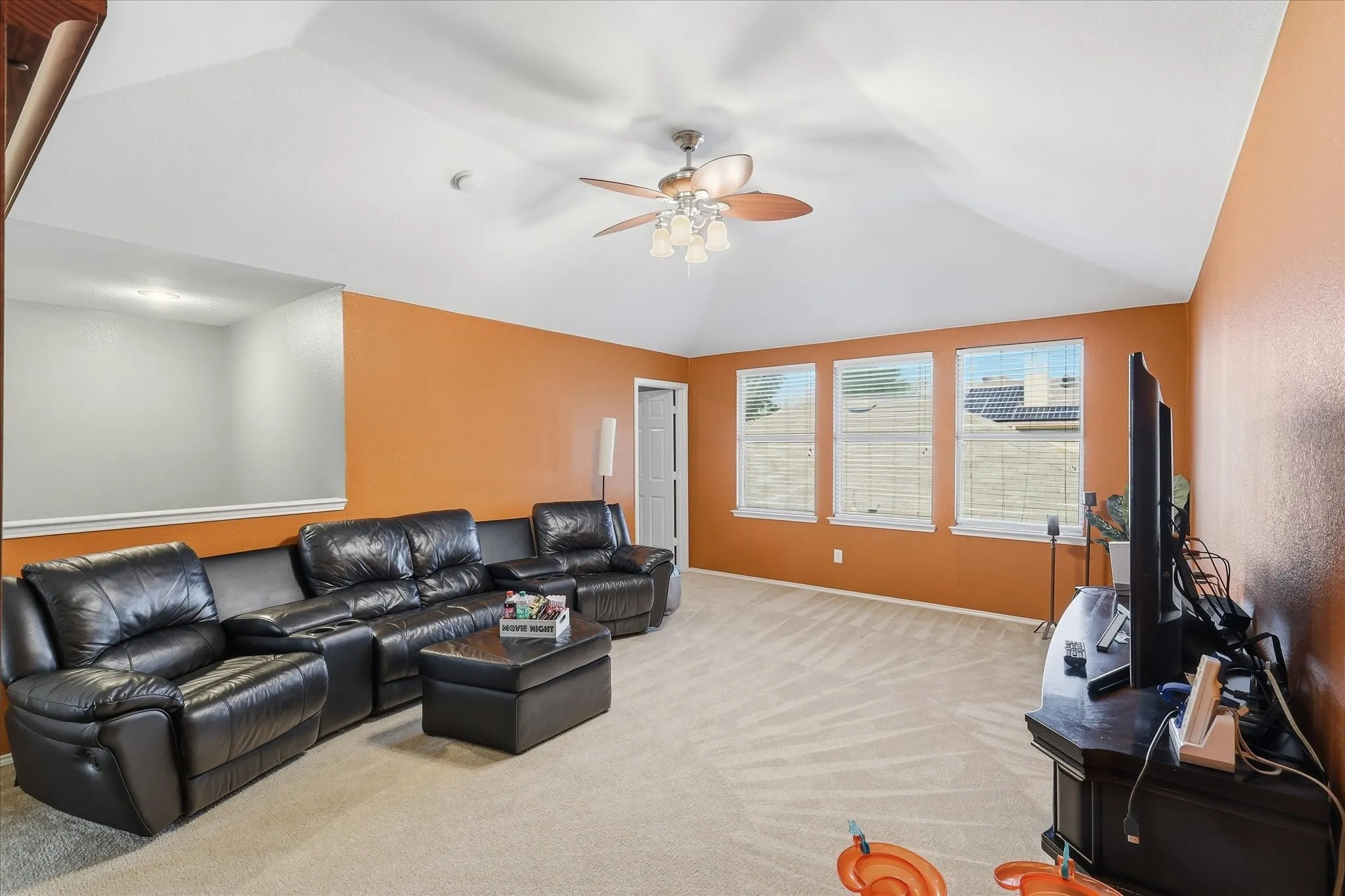 Spacious room featuring vaulted ceilings, a ceiling fan with integrated lighting, three windows with blinds, and light-toned carpet flooring