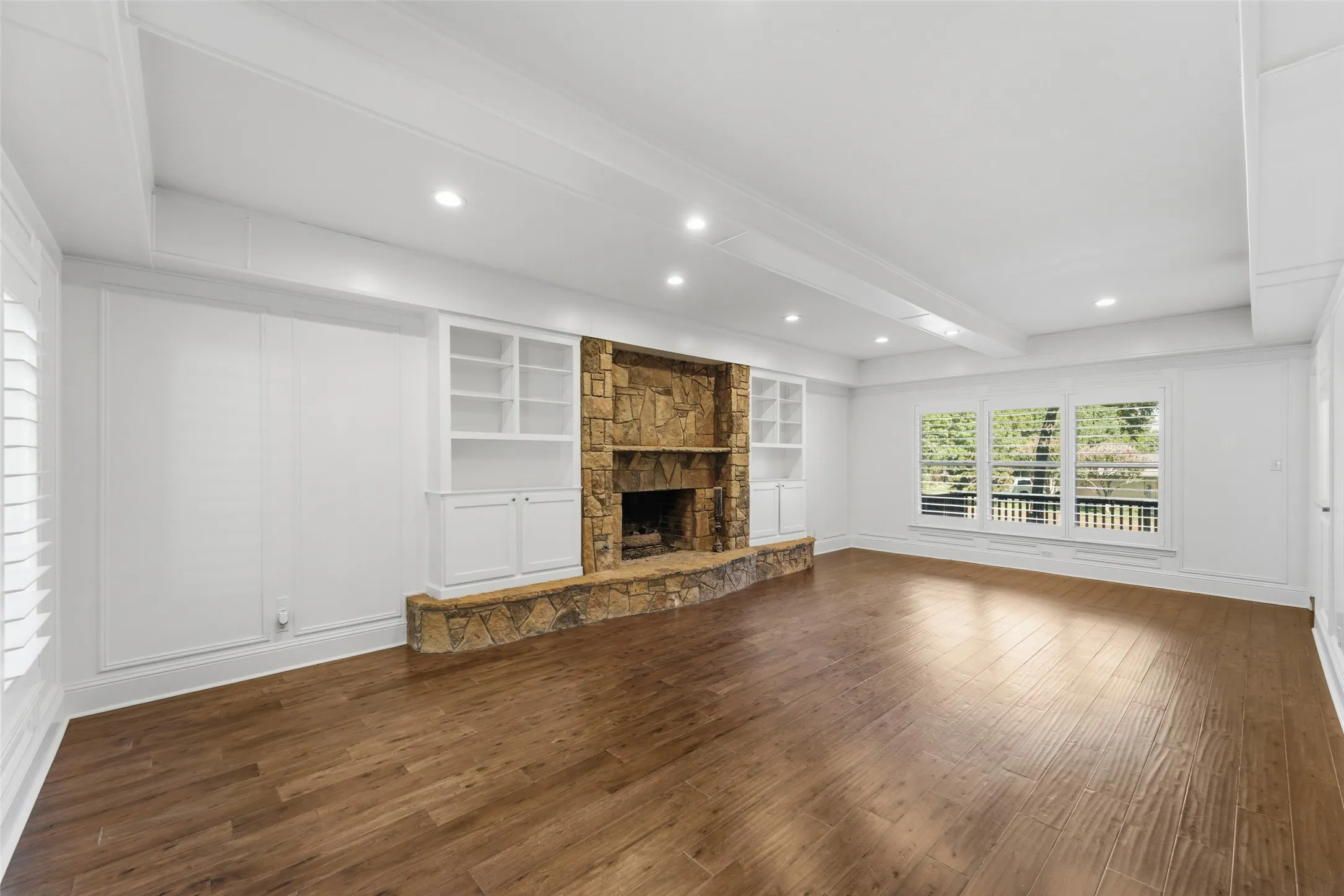 Spacious room featuring wood-finish flooring, a stone fireplace, built-in shelving, recessed lighting, and large windows with plantation shutters