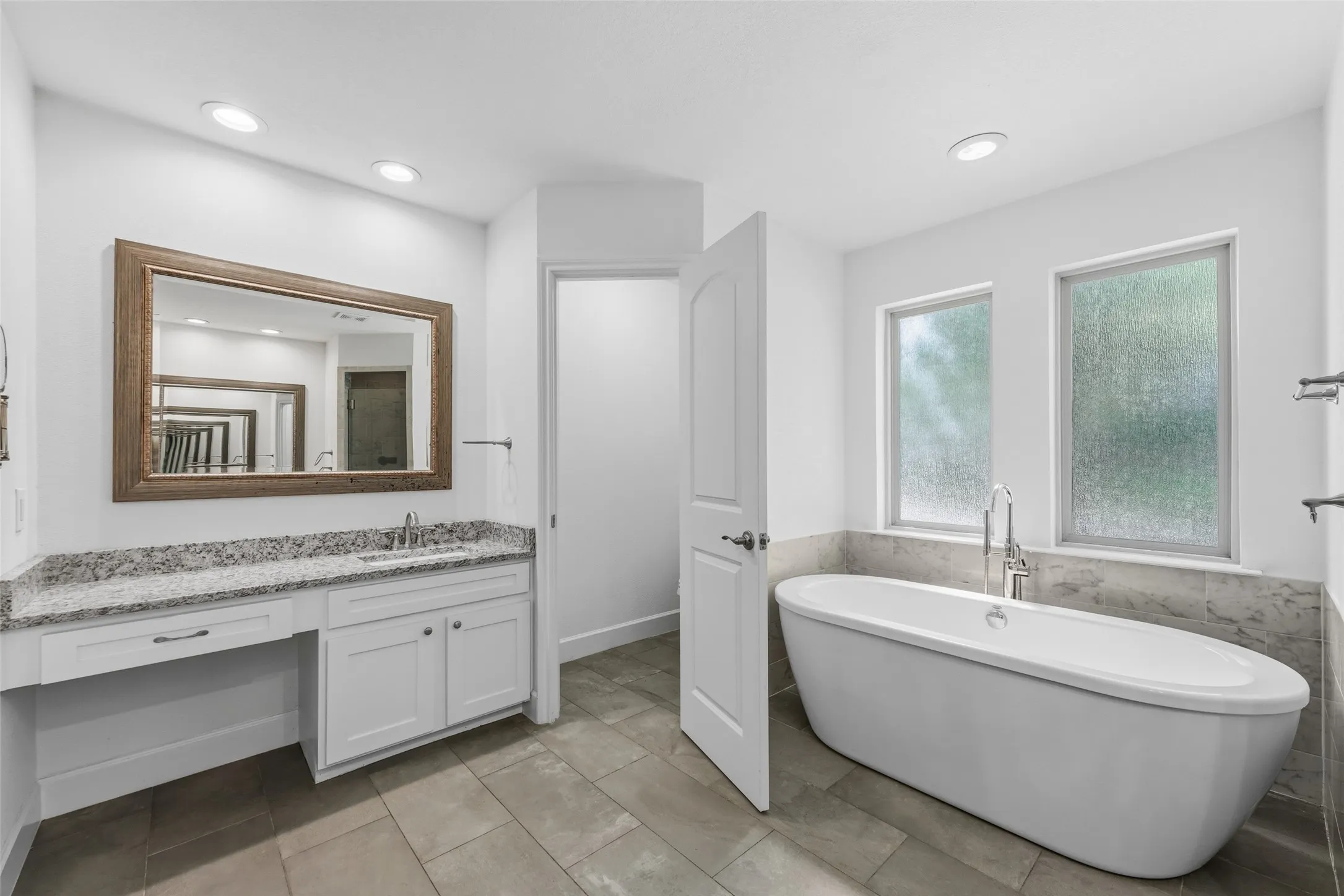Spacious bathroom featuring a freestanding soaking tub, dual windows, a white vanity with granite-style countertop, and rectangular floor tiling