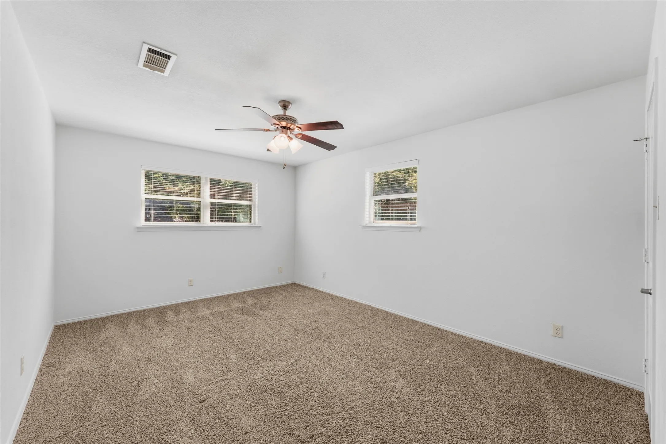 Spacious interior room featuring light brown carpeting, white painted walls, two windows with horizontal blinds, and a ceiling fan with integrated lighting