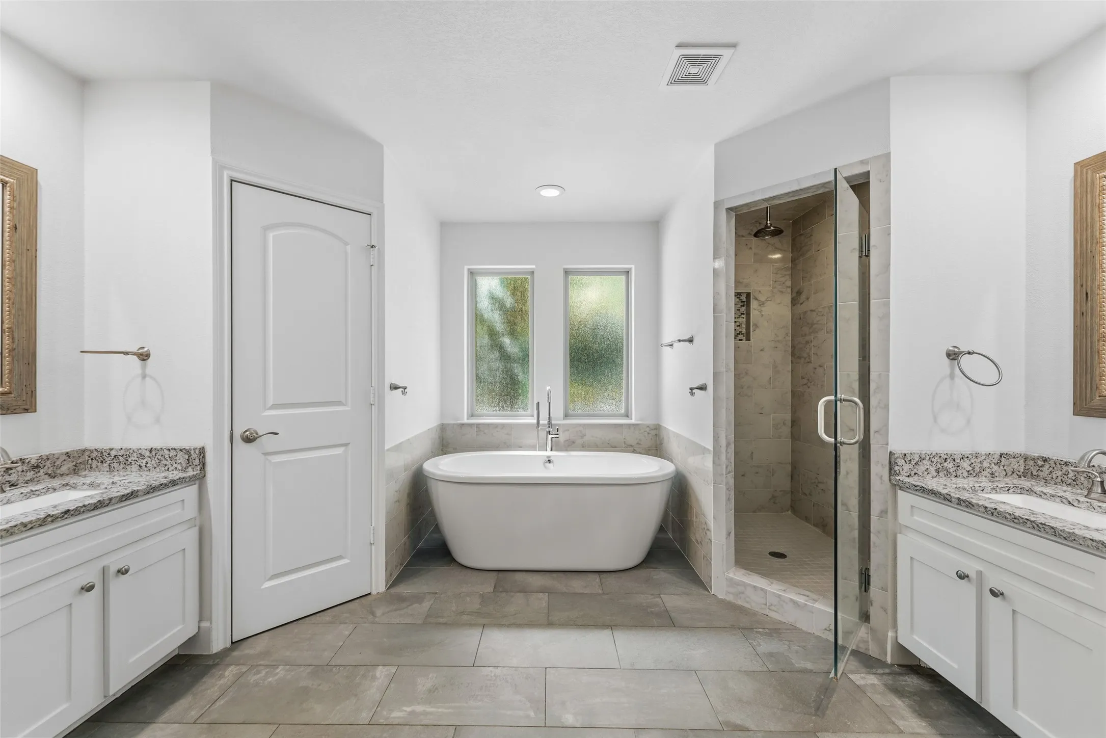 Contemporary bathroom featuring a freestanding tub, a glass-enclosed shower, dual vanities with granite countertops, and light-toned tile flooring