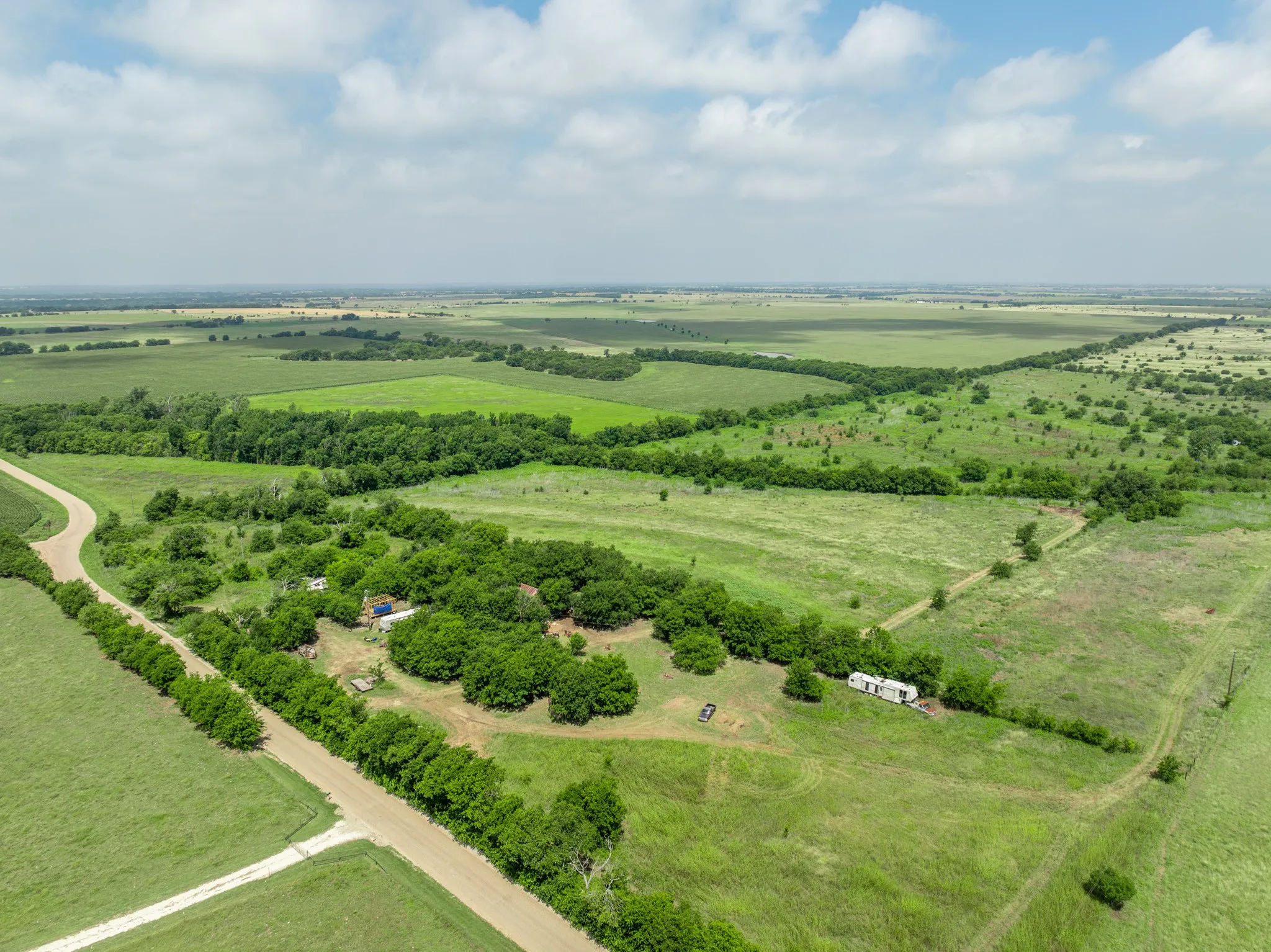 Expansive rural landscape featuring a dirt road, open fields, scattered trees, and distant agricultural areas