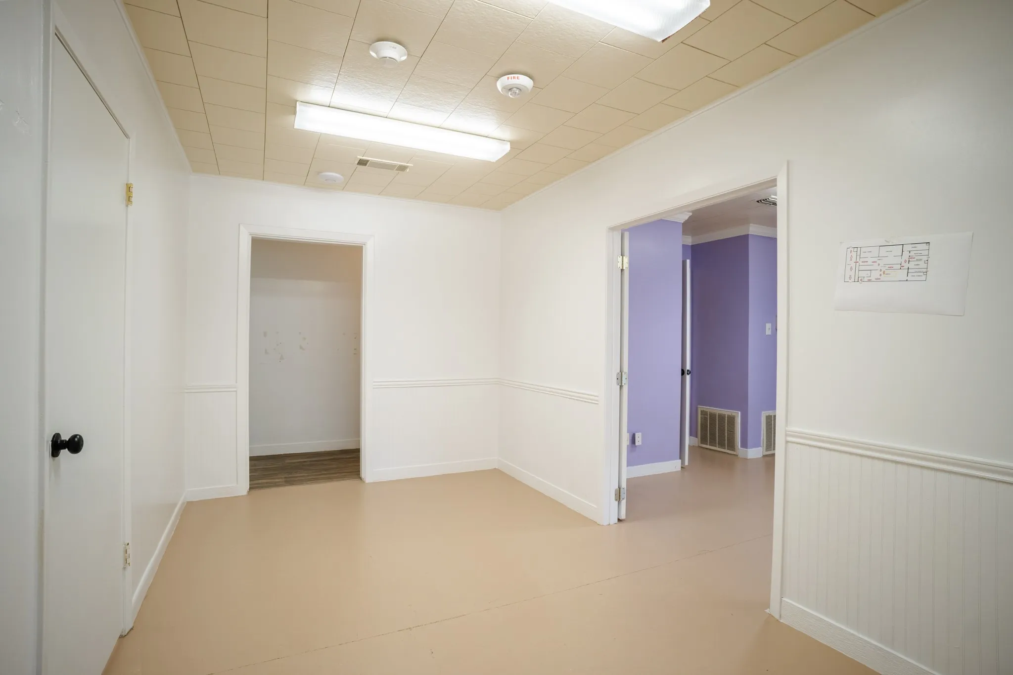 Room featuring light-toned flooring, white walls with wainscoting, and a drop ceiling with integrated fluorescent lighting
