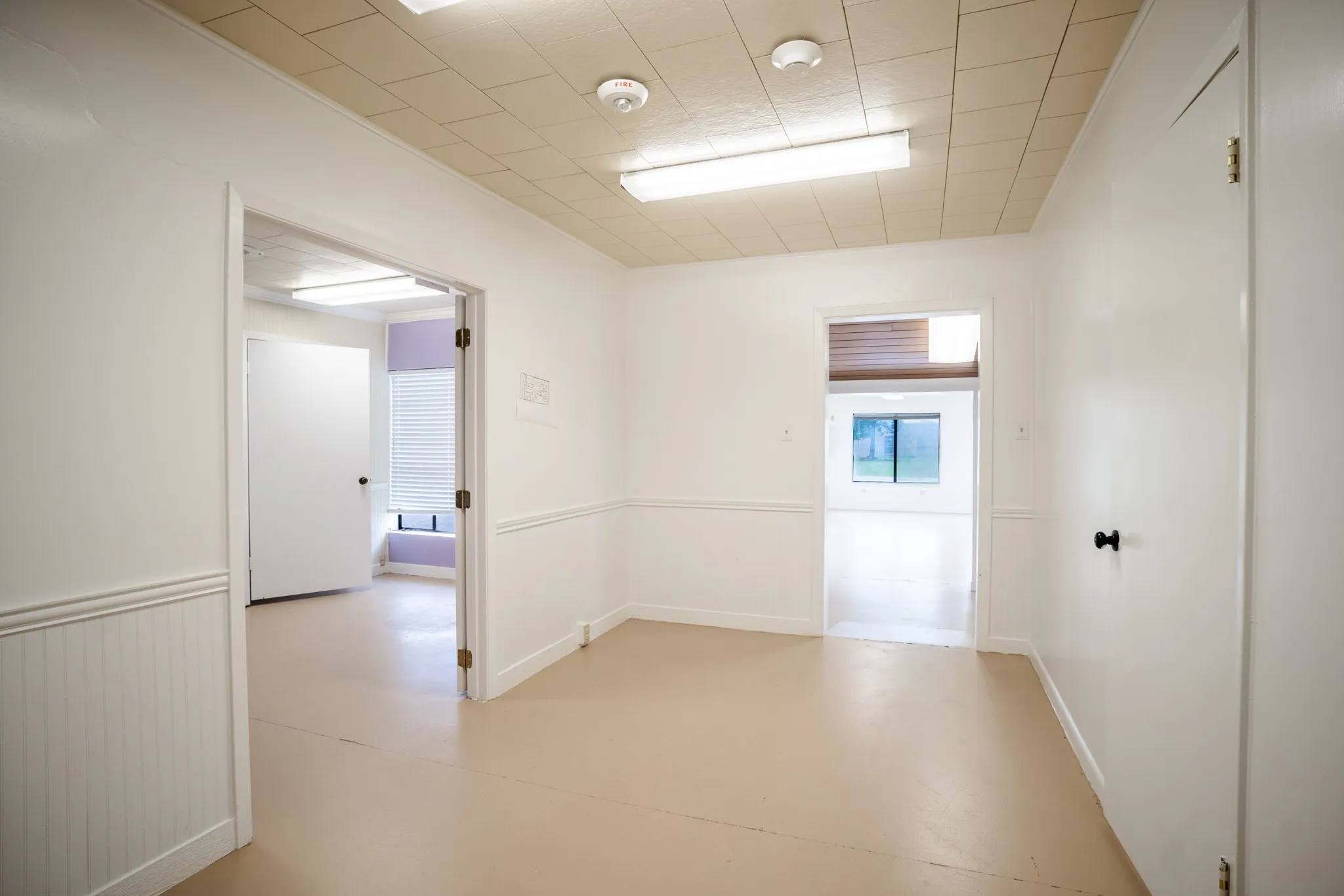 Spacious interior room with light-toned flooring, white walls featuring beadboard wainscoting and chair rail molding, and a grid-pattern drop ceiling with fluorescent lighting