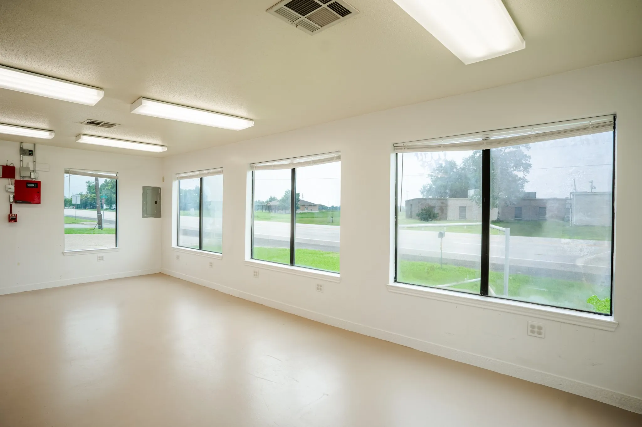 Spacious room featuring multiple large windows, light-colored flooring, white walls, and overhead fluorescent lighting