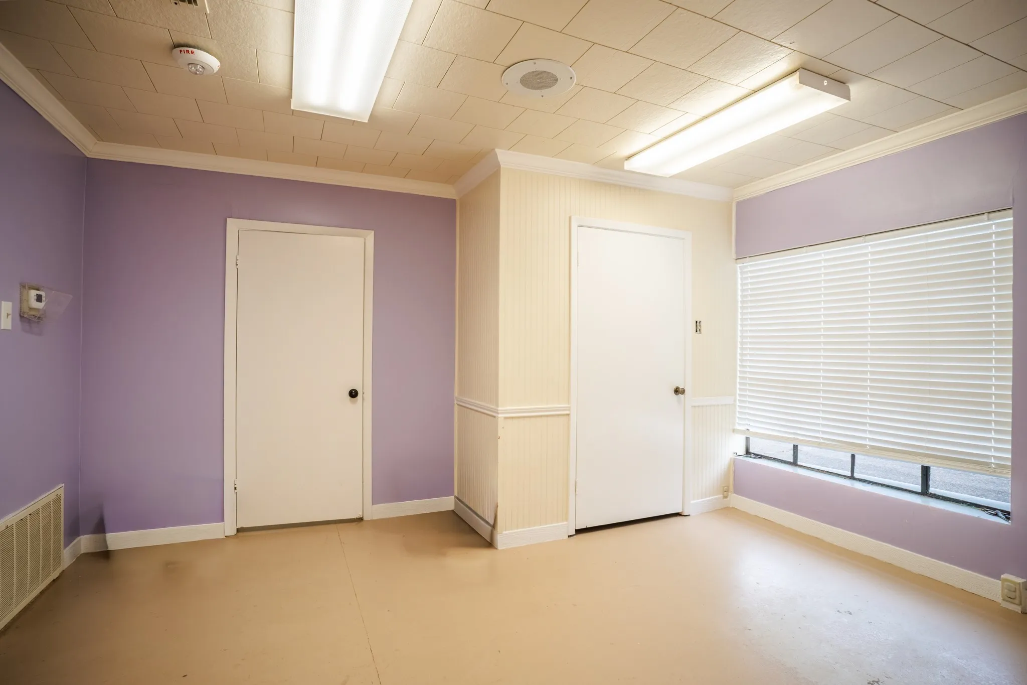 Interior space featuring a window with horizontal blinds, two white doors, perimeter baseboards, and overhead fluorescent lighting fixtures
