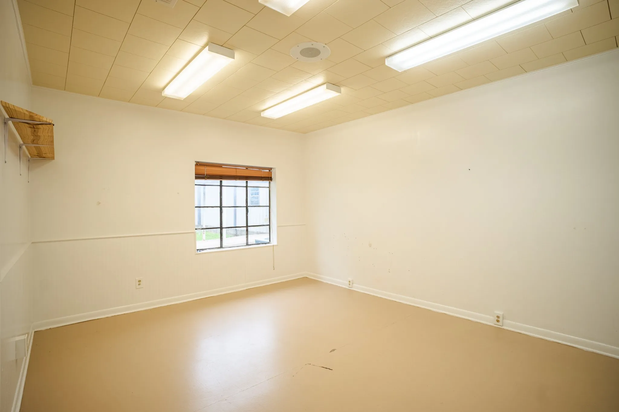 Room featuring a grid-pane window with wood-finish blinds, light-colored walls with wainscoting, a drop panel ceiling with overhead fluorescent lighting, and a beige floor