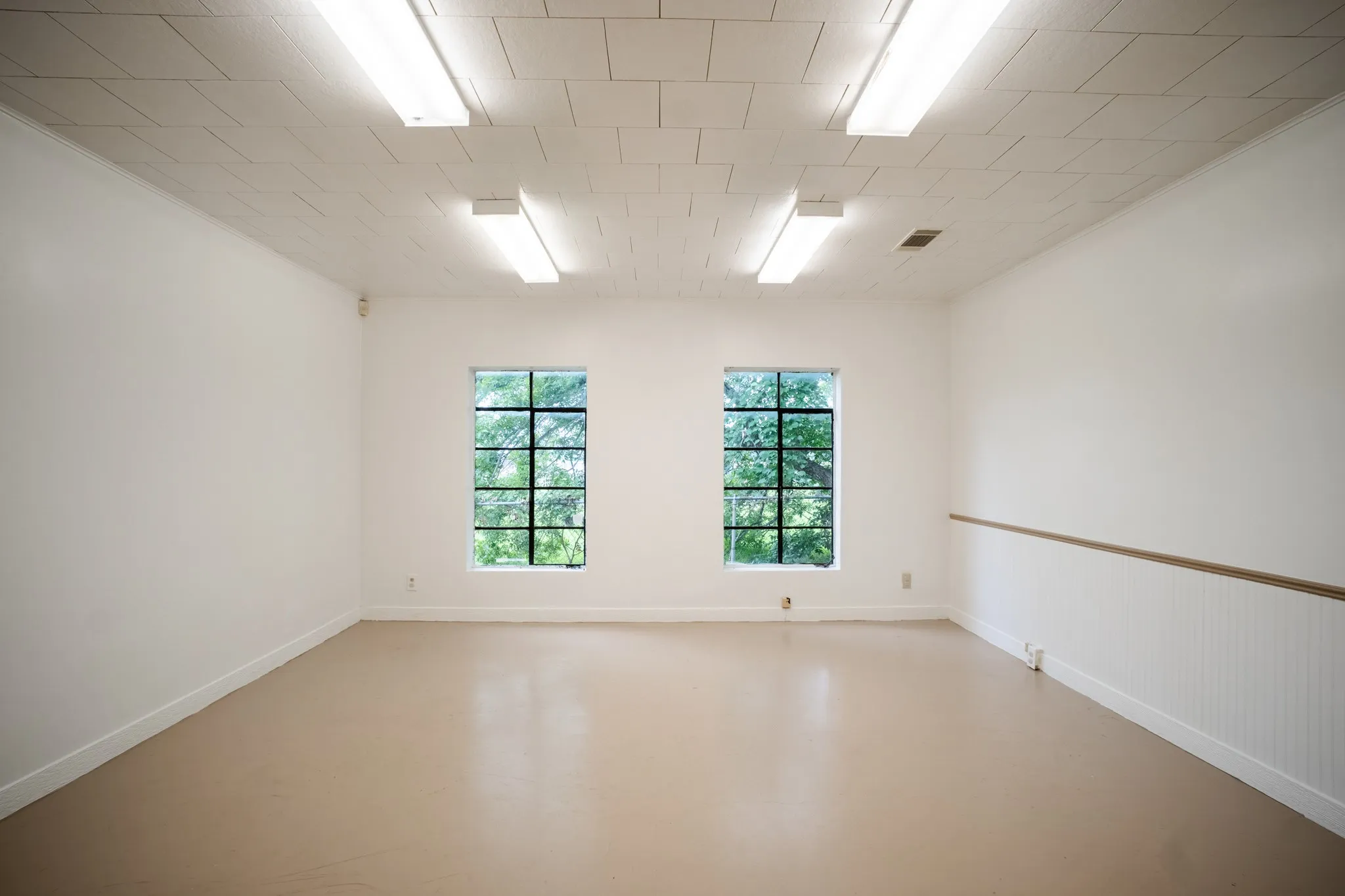 Spacious room featuring two large grid-pattern windows, beadboard wainscoting with a wooden rail, recessed fluorescent lighting, and light-toned flooring