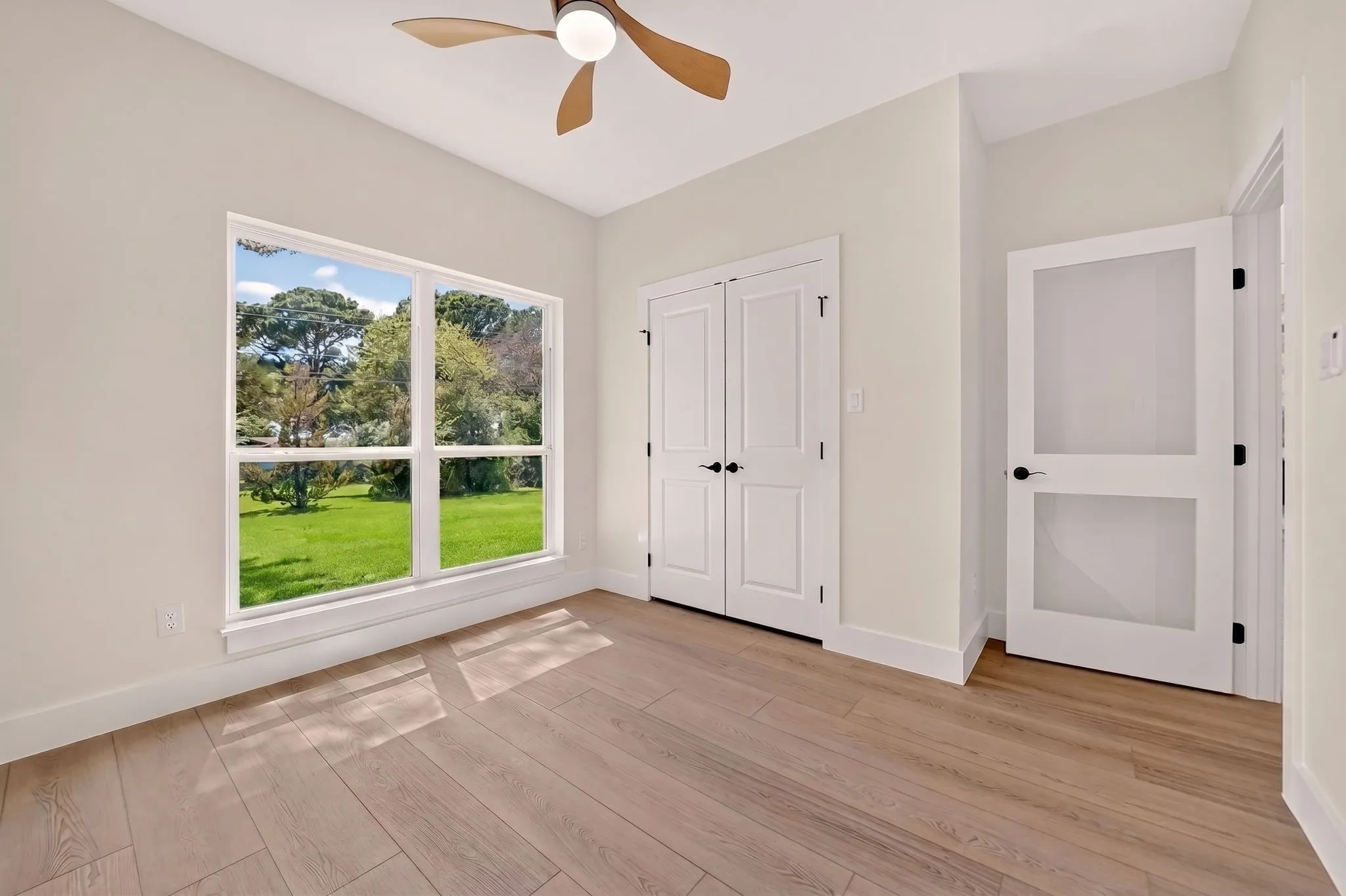 Expansive window, wood-finish flooring, ceiling fan, double closet doors with black hardware, and an interior door with frosted glass panels