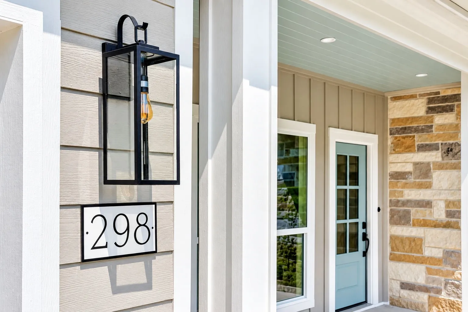 Board and batten siding, decorative stone facade, light blue entry door, white trim windows, and a black framed outdoor lantern