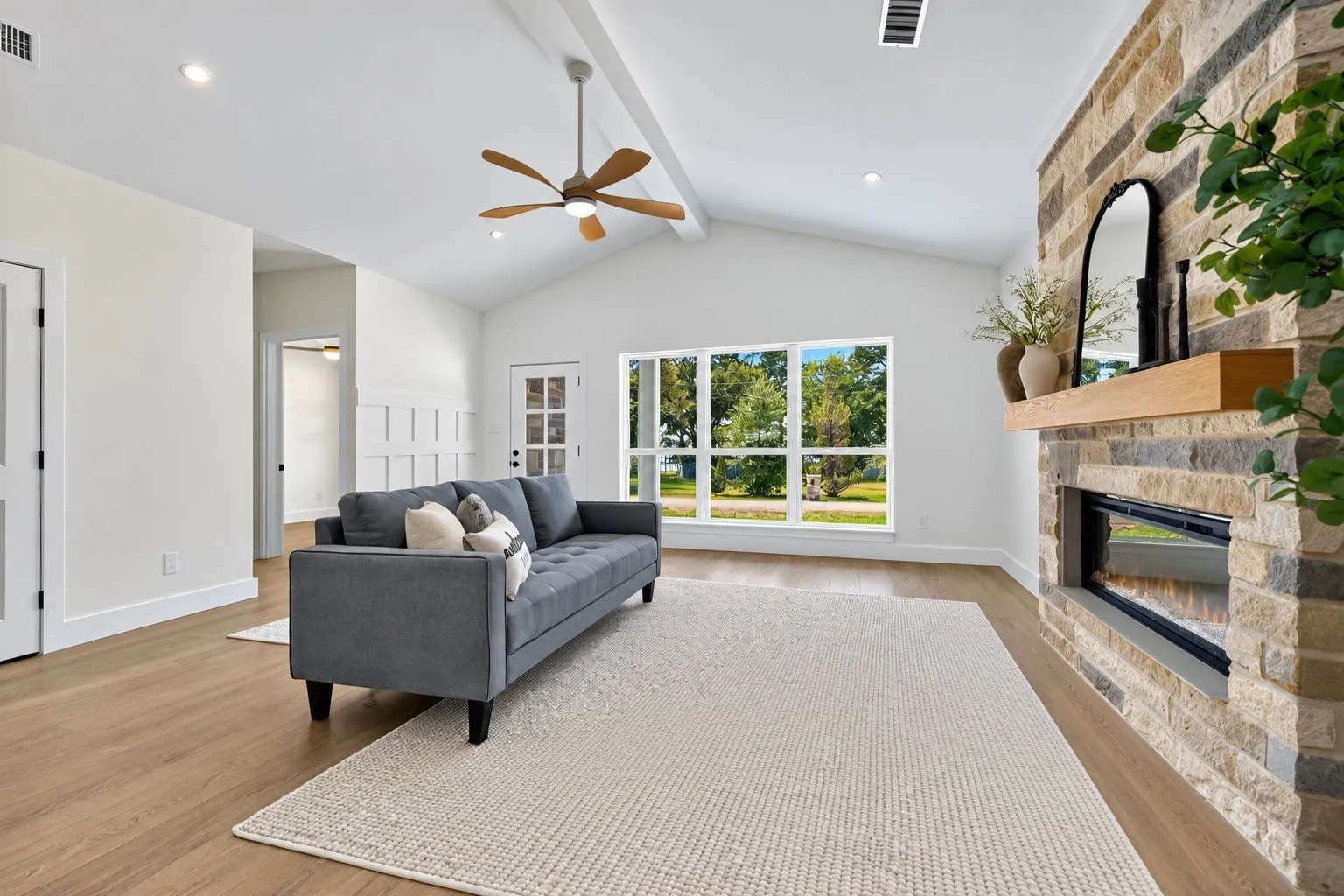 Vaulted ceilings, wood-finish flooring, stone-clad fireplace with wood mantel, large pane windows, and a ceiling fan with light