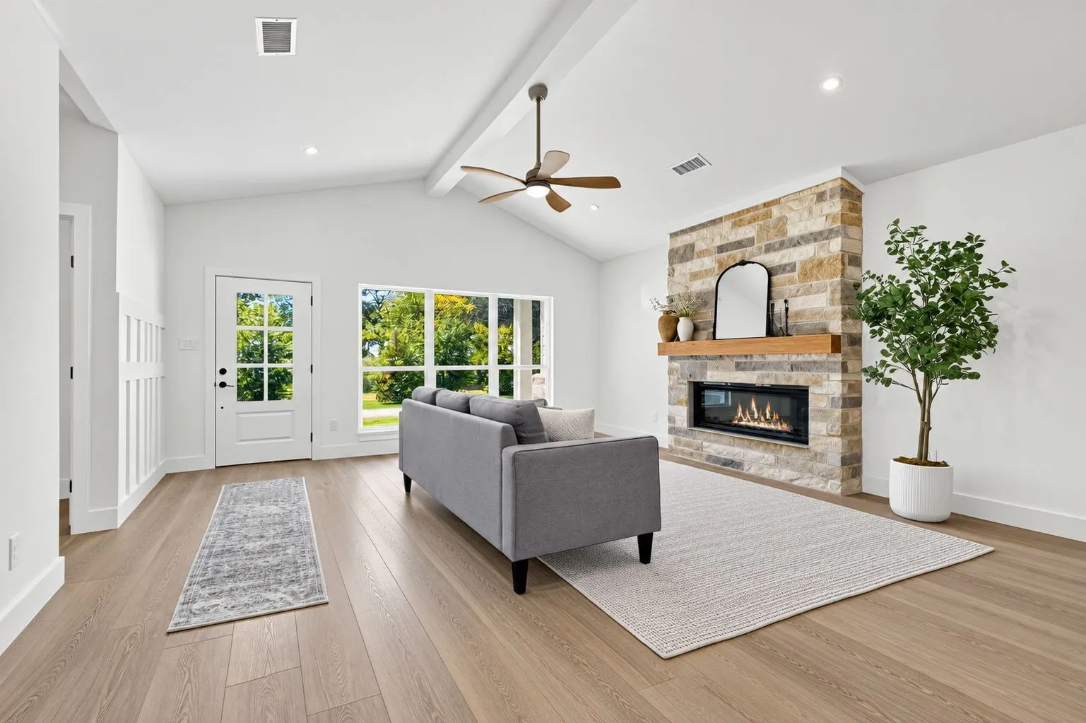 Vaulted ceiling with exposed beam, stone fireplace with wood mantle, modern linear gas fireplace, wood-finish flooring, and large window