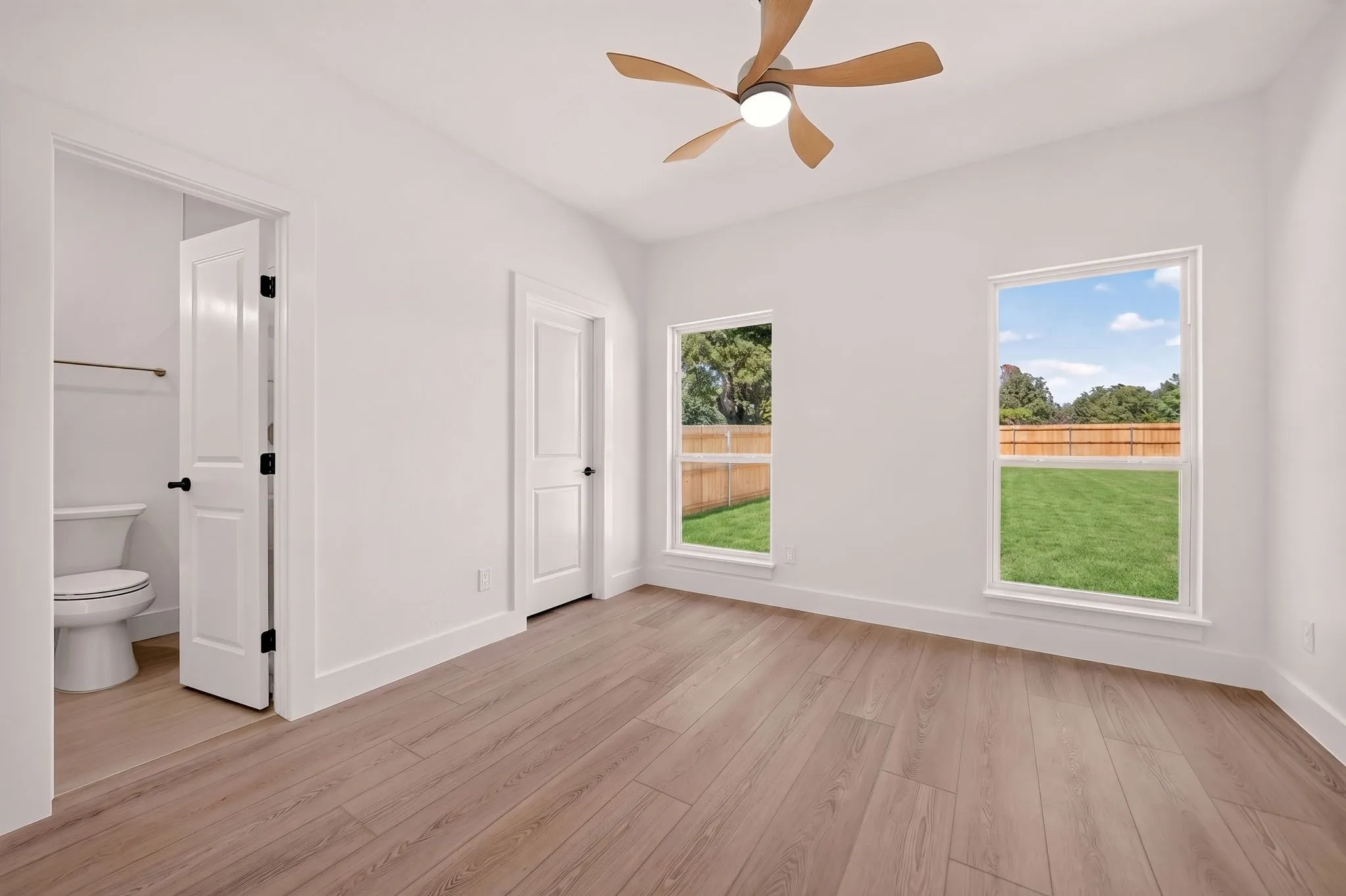 Wood-finish flooring, two large windows, modern ceiling fan, white paneled doors, and an attached private toilet room