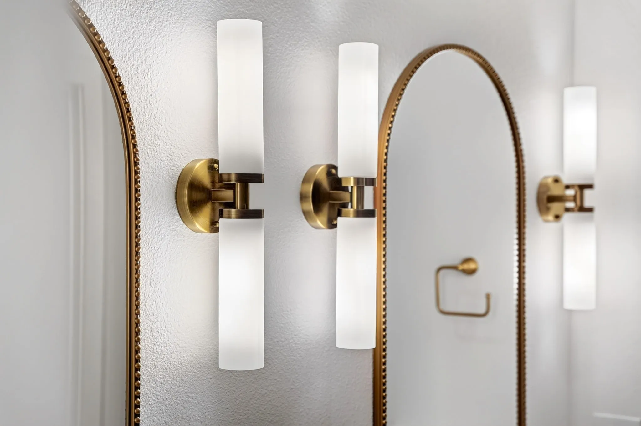 Brushed brass wall sconces featuring frosted glass cylinders and an arched mirror with a beaded frame
