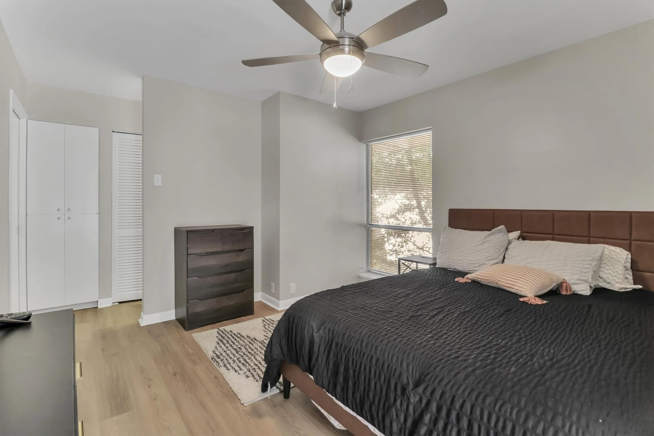 Spacious bedroom featuring wood-finish flooring, a contemporary ceiling fan with integrated lighting, a large window providing ample natural light, and built-in white cabinetry with louvered doors