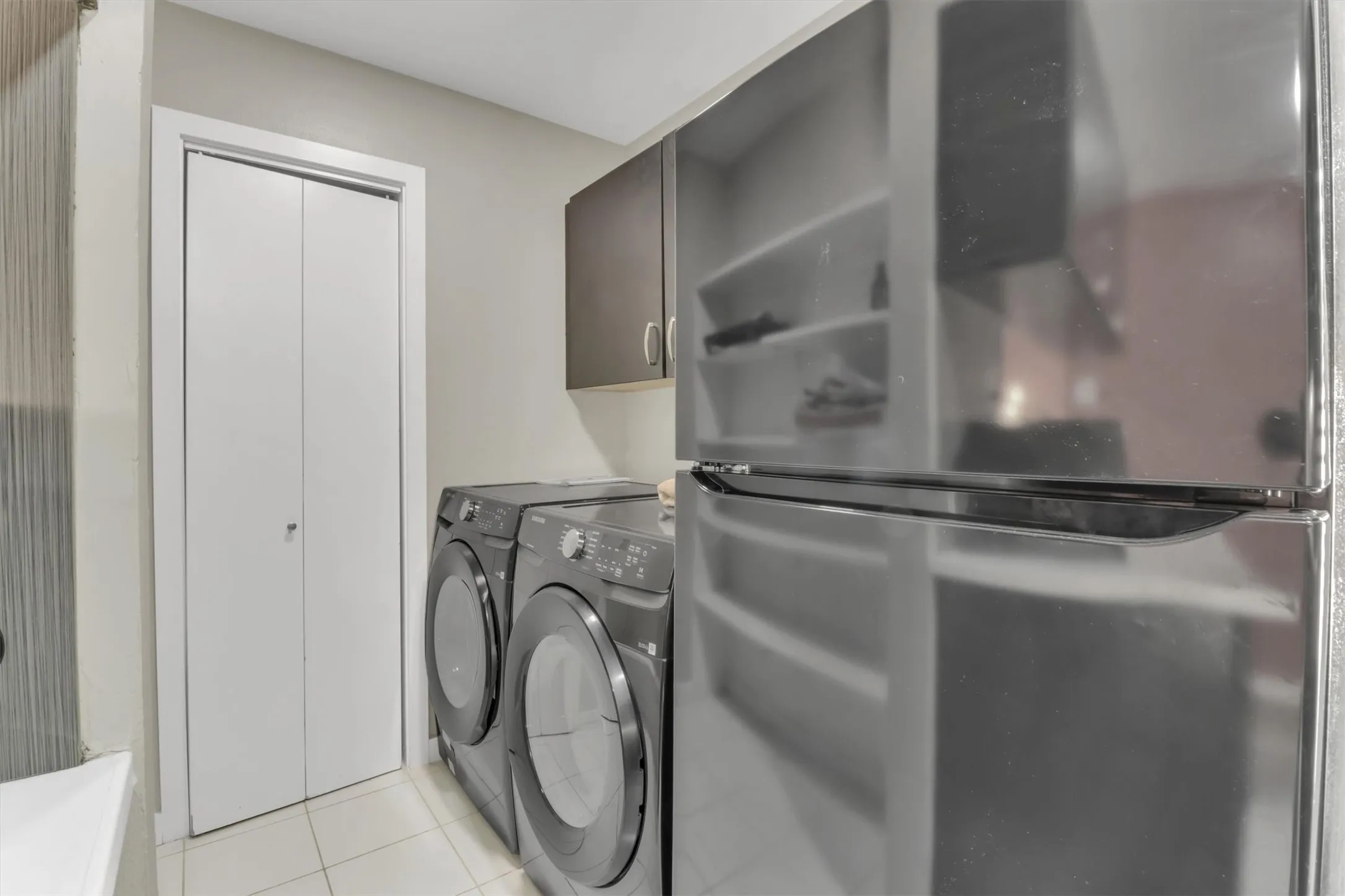 Utility area featuring tiled flooring, a built-in pantry with bi-fold doors, stacked washer and dryer units, a dark-toned wall cabinet, and an adjacent refrigerator
