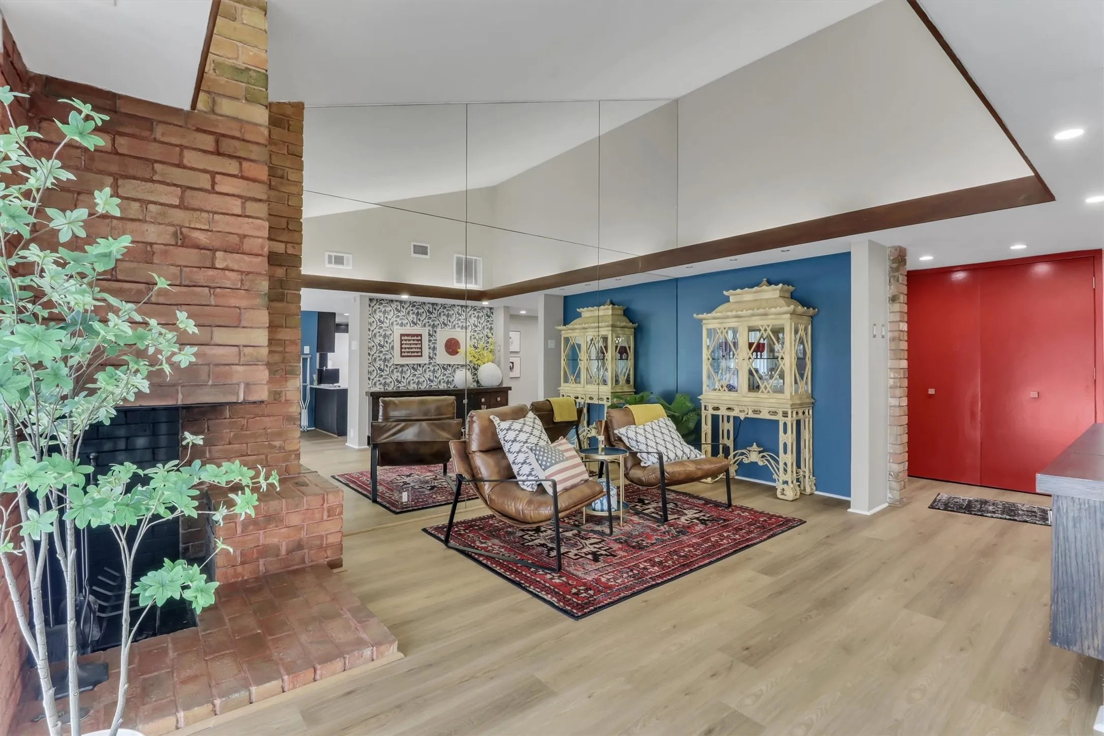 Exposed brick fireplace with a raised hearth, light wood-finish flooring, and a vibrant red entry door