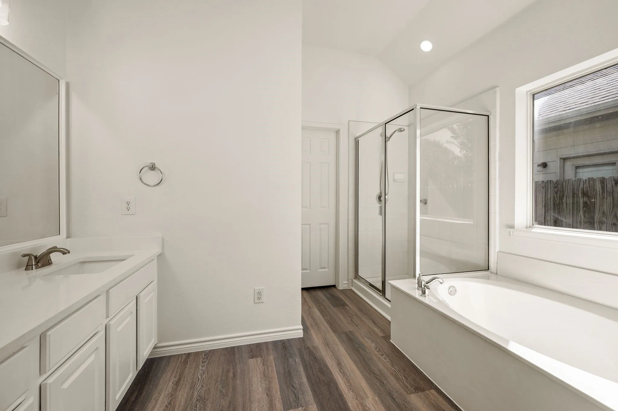 Spa-inspired primary bathroom featuring dual vanities with white cabinetry, a soaking tub with tile surround, a frameless glass-enclosed shower, and wood-finish flooring throughout the space