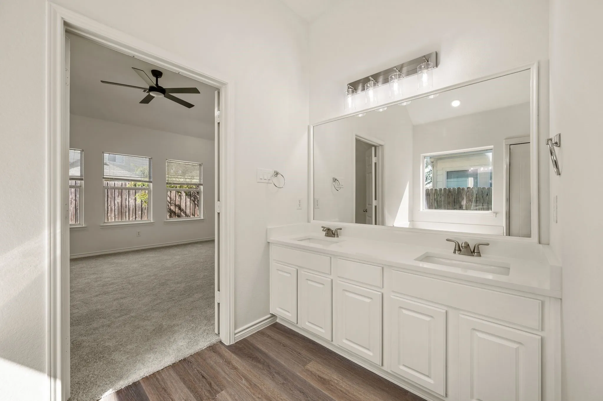 Spa-inspired primary bathroom featuring dual vanities with white cabinetry, a large framed mirror with integrated lighting, and wood-finish flooring