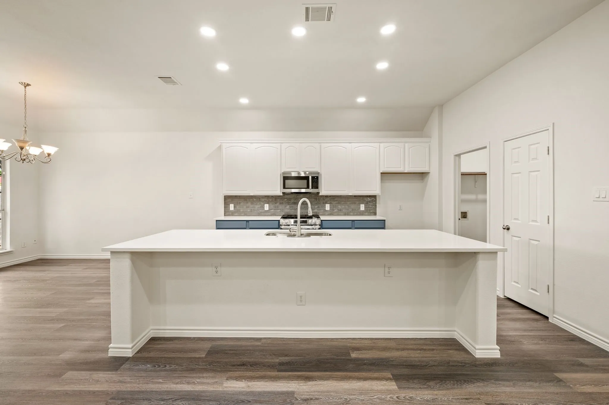 Spacious open-concept kitchen featuring a large waterfall-edge island with white quartz countertop, stainless steel appliances, gray tile backsplash, white cabinetry, recessed ceiling lighting, and wood-finish flooring throughout the adjoining living areas
