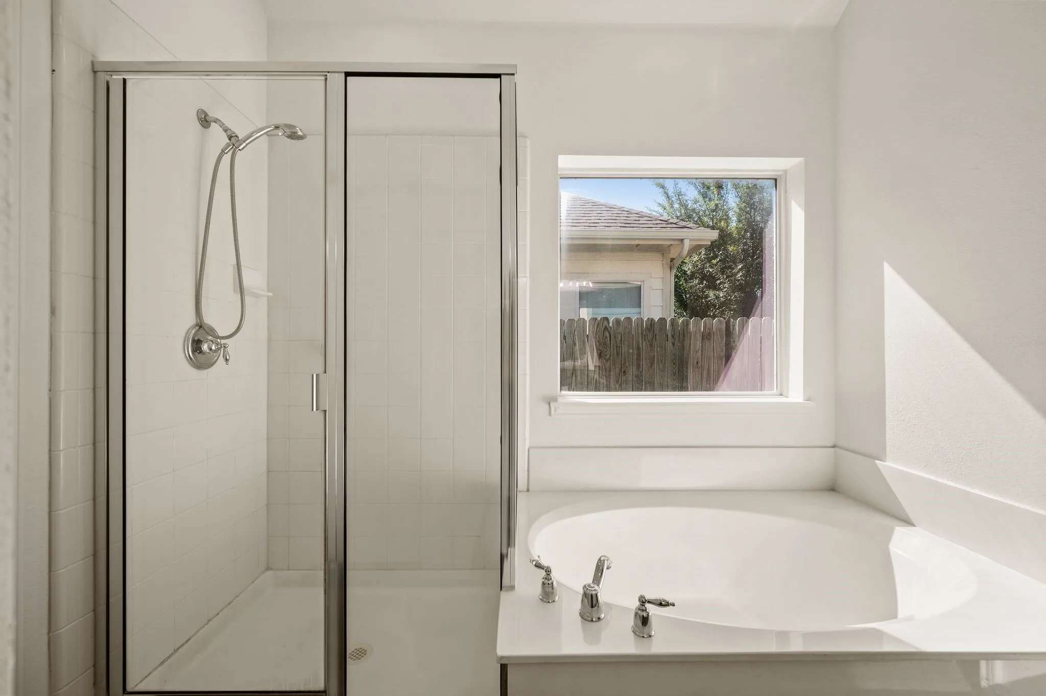 Modern primary bathroom featuring a frameless glass-enclosed shower with chrome fixtures, a soaking tub with deck-mounted faucet and handheld showerhead, white subway tile surround, and natural light from a casement window overlooking the exterior grounds
