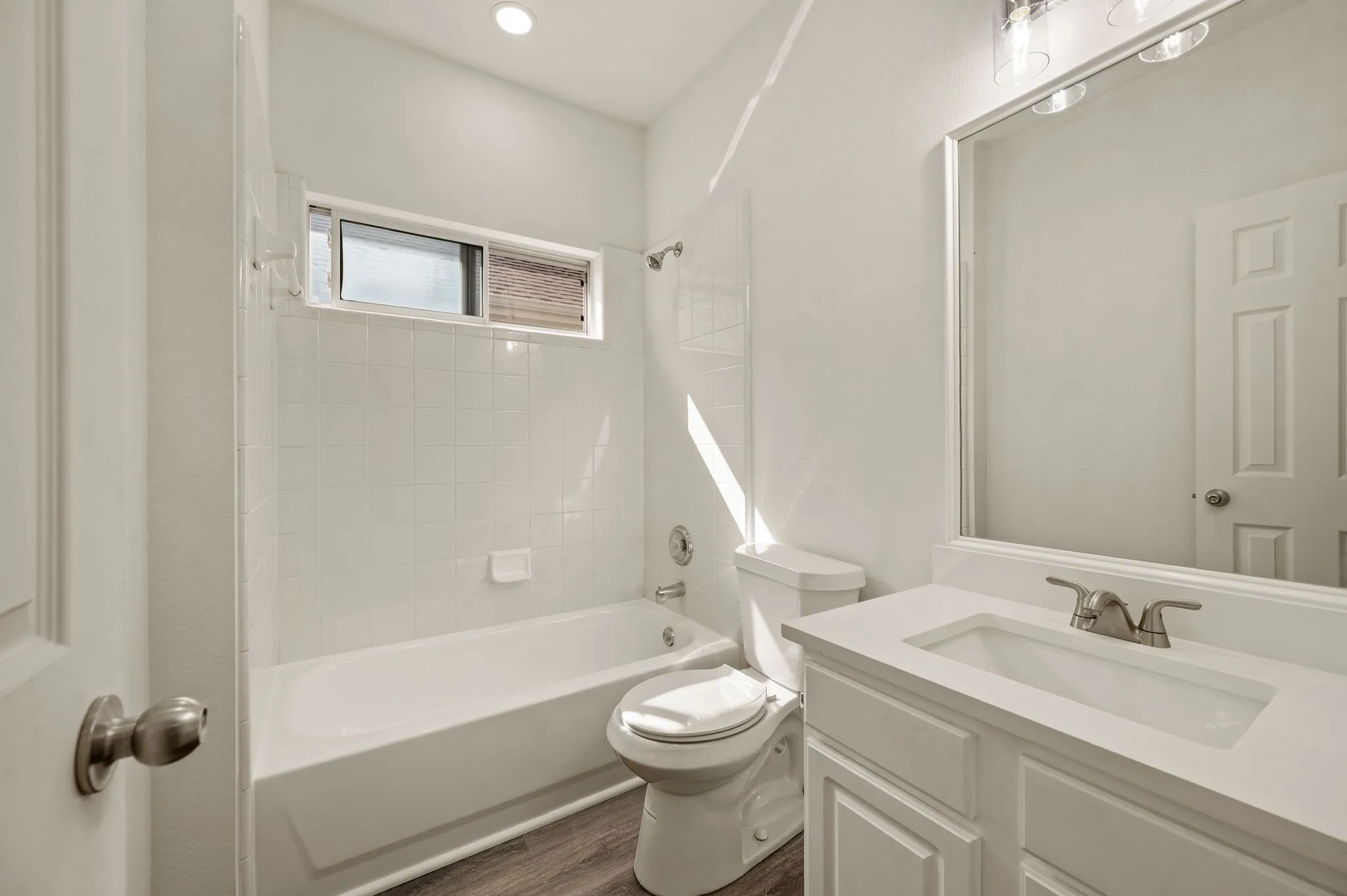 Bright, updated bathroom featuring a white subway tile surround, integrated soaking tub with glass enclosure, dual vanity with rectangular sink and brushed nickel faucet, ceramic toilet, wood-finish flooring, and recessed ceiling lighting with a transom window for natural illumination