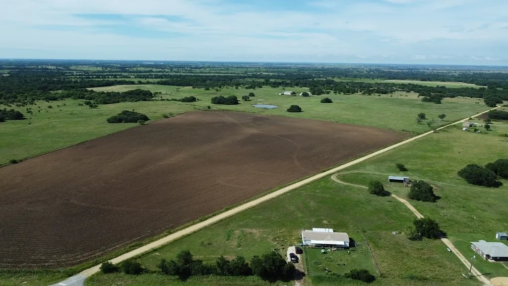 Expansive rural landscape next to a large cultivated field, open grassy areas, scattered mature trees, and a visible dirt road