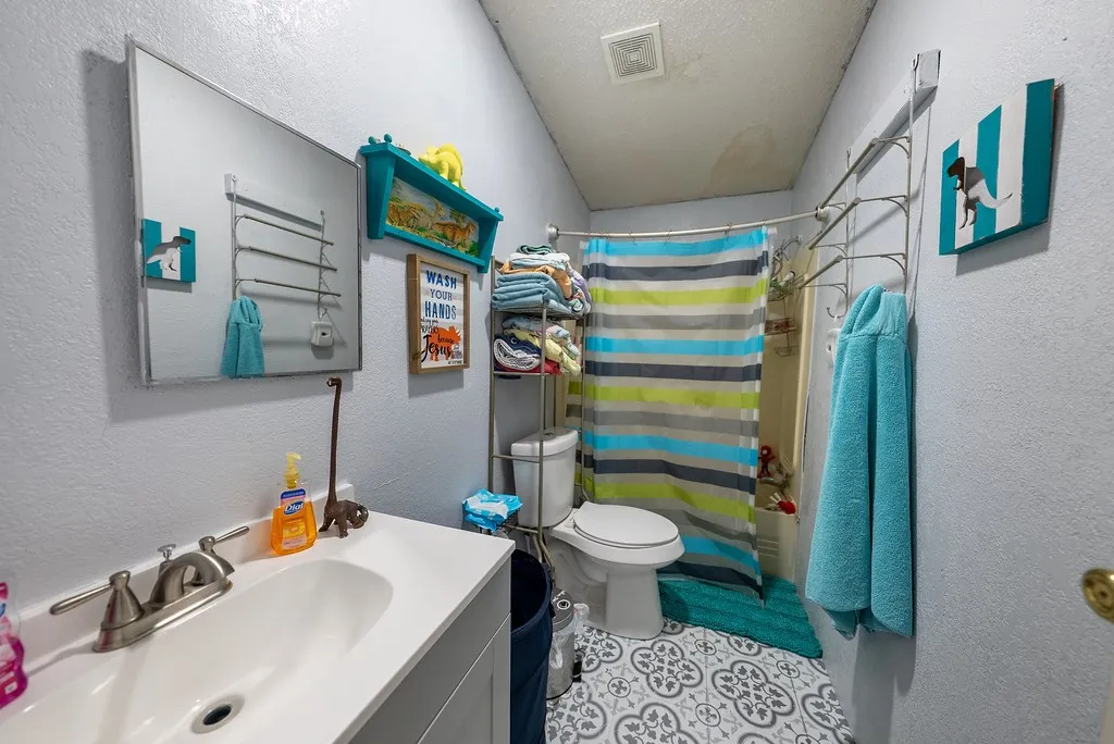 Bathroom featuring a single-sink vanity with brushed nickel fixtures, a wall-mounted mirror, patterned floor tiles, a commode, and a shower area