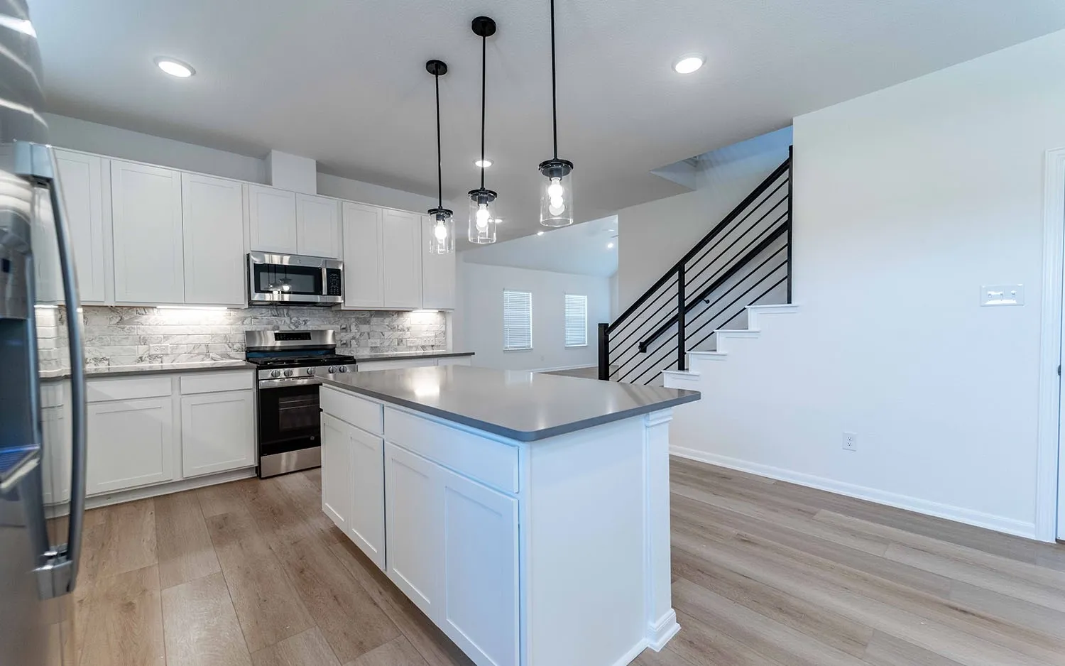Modern kitchen featuring white shaker cabinetry, stainless steel appliances, a light-toned wood-finish floor, an island with a light countertop, and contemporary pendant lighting
