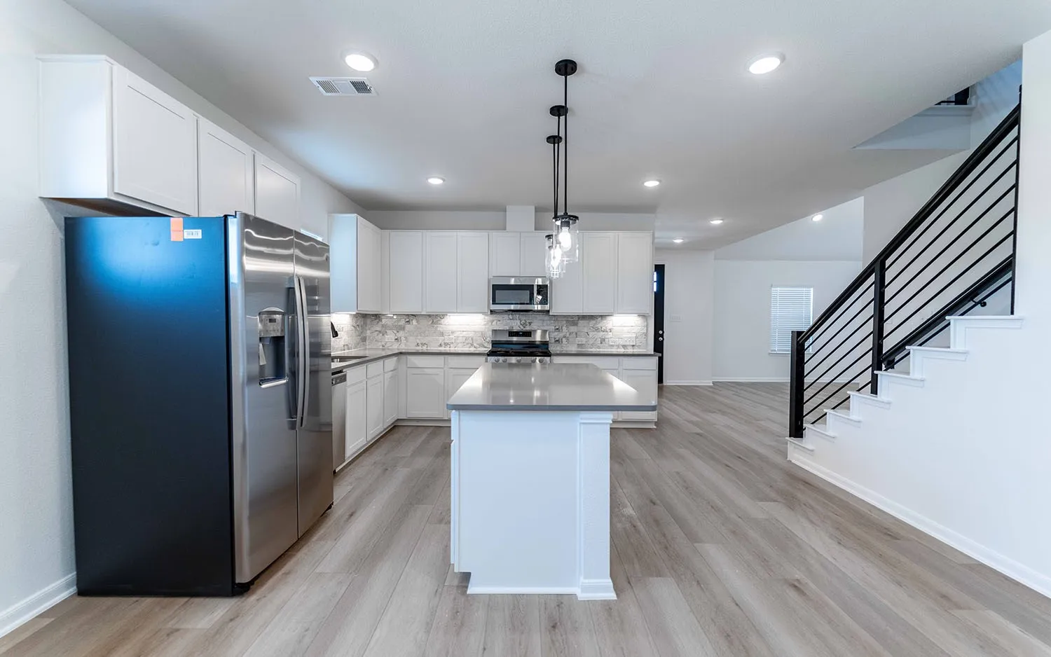 Open-concept living space featuring wood-finish flooring, a kitchen island with a light-toned countertop, white cabinetry, stainless steel appliances, and a staircase with a black metal railing