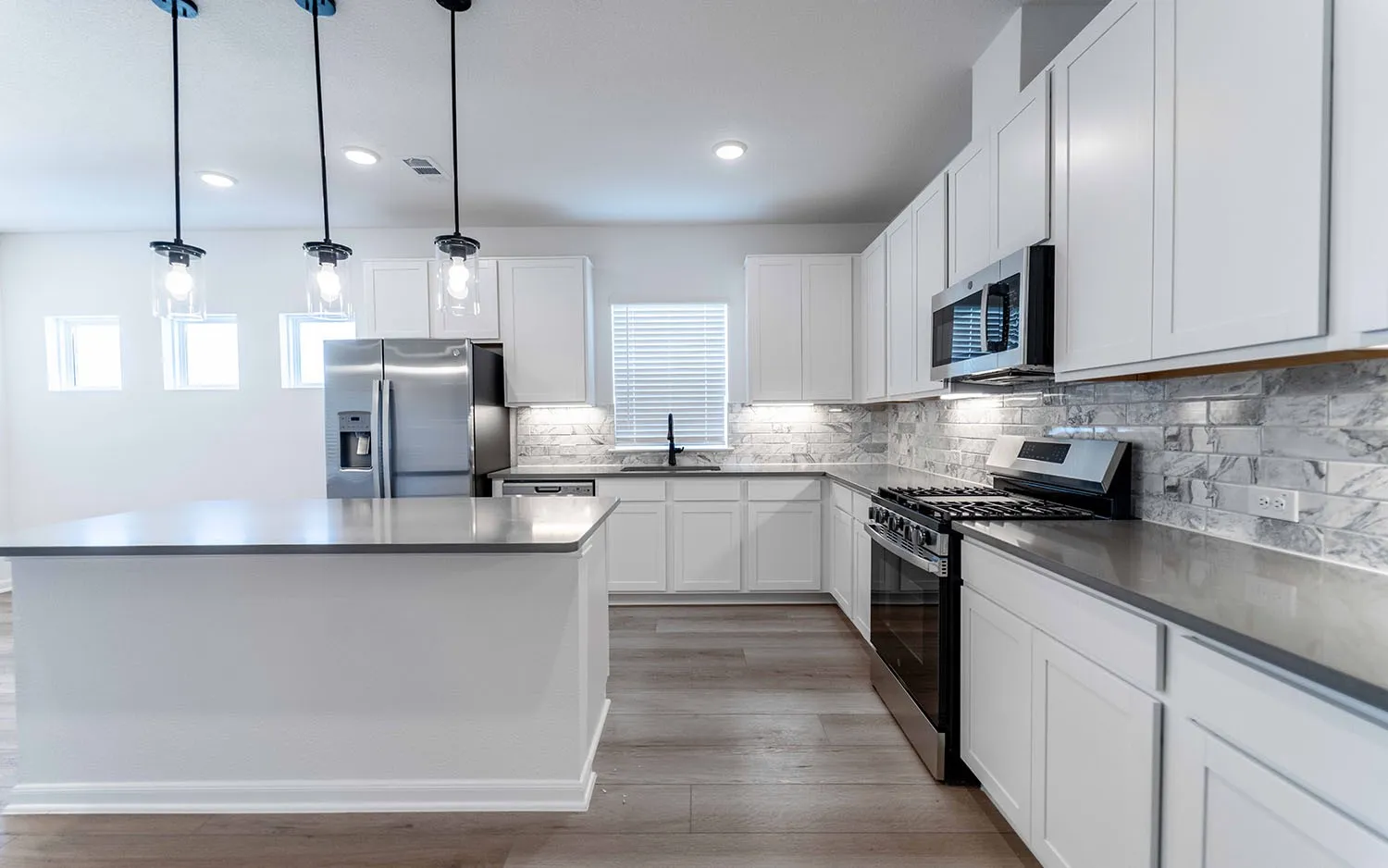 Spacious kitchen featuring an expansive center island with a dark countertop, white shaker-style cabinetry, stainless steel appliances, a grey and white subway tile backsplash, and wood-finish flooring
