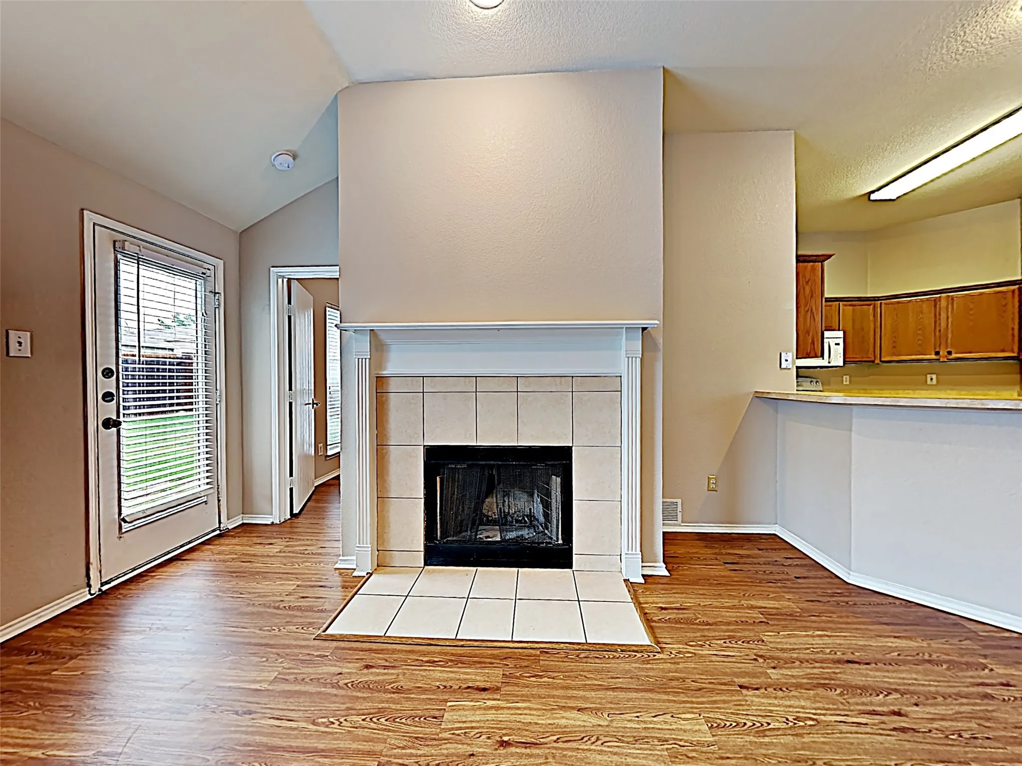 Living area featuring wood-finish flooring, a tile-surround fireplace with a white mantel, and a door with integrated blinds opening to the exterior