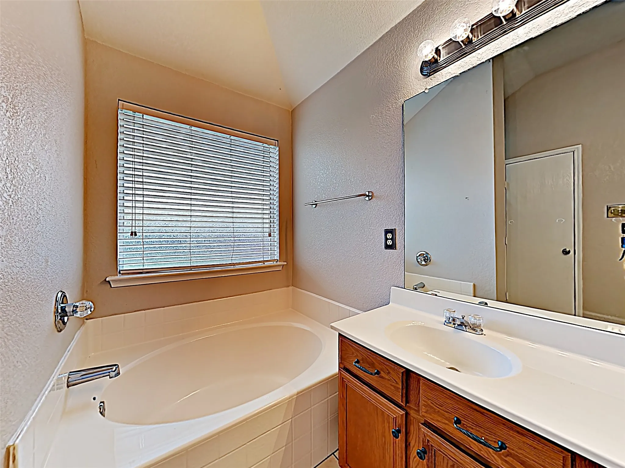 Bathroom featuring a built-in tub, a single vanity with wood-finish cabinetry, a framed mirror, and a window with blinds
