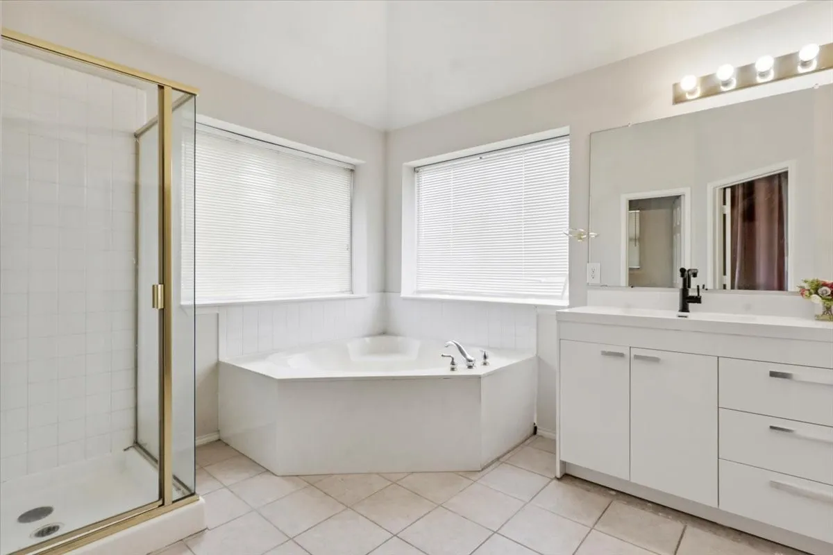 Bathroom featuring a corner soaking tub, a glass-enclosed shower with gold-tone framing, a double window with blinds, and a white vanity with an integrated sink and large mirror
