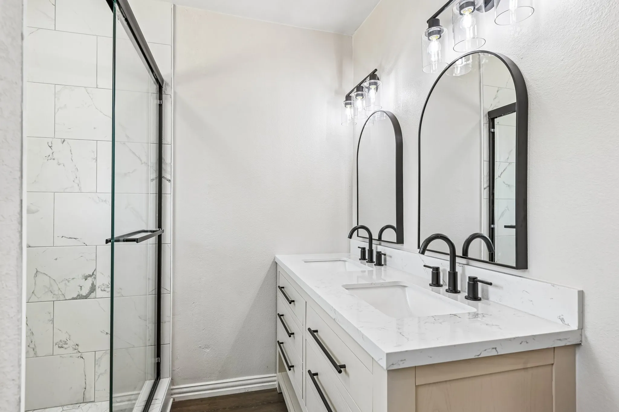 Bathroom featuring a glass-enclosed shower with white tiled walls, a double vanity with white countertops, and dual arched mirrors with matte black frames