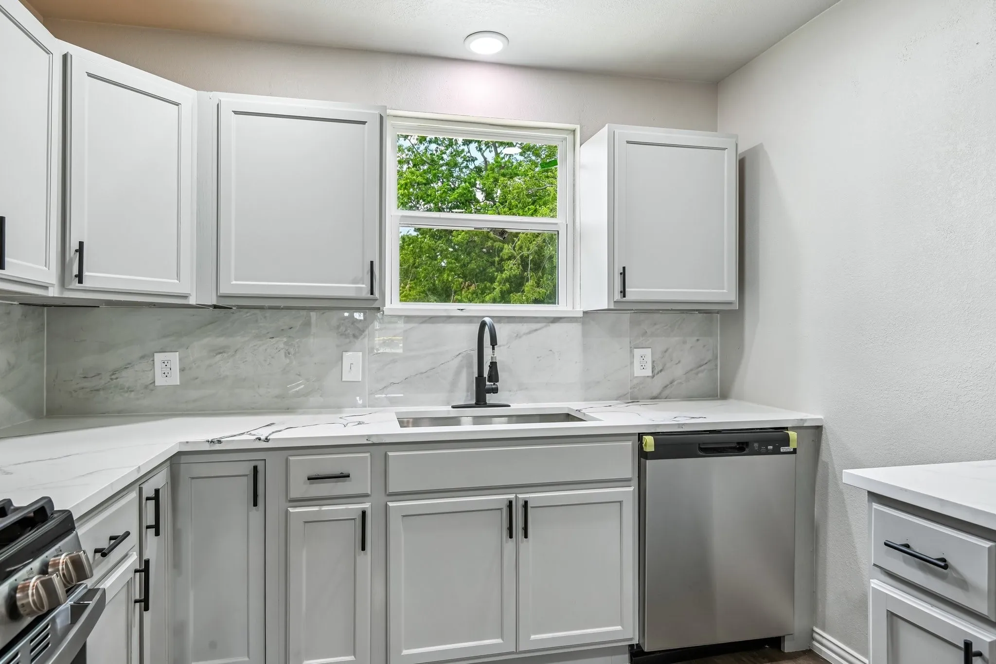 Modern kitchen featuring light gray cabinetry with black hardware, white countertops with subtle veining, a stainless steel sink with a black gooseneck faucet, a stainless steel dishwasher, and a window above the sink