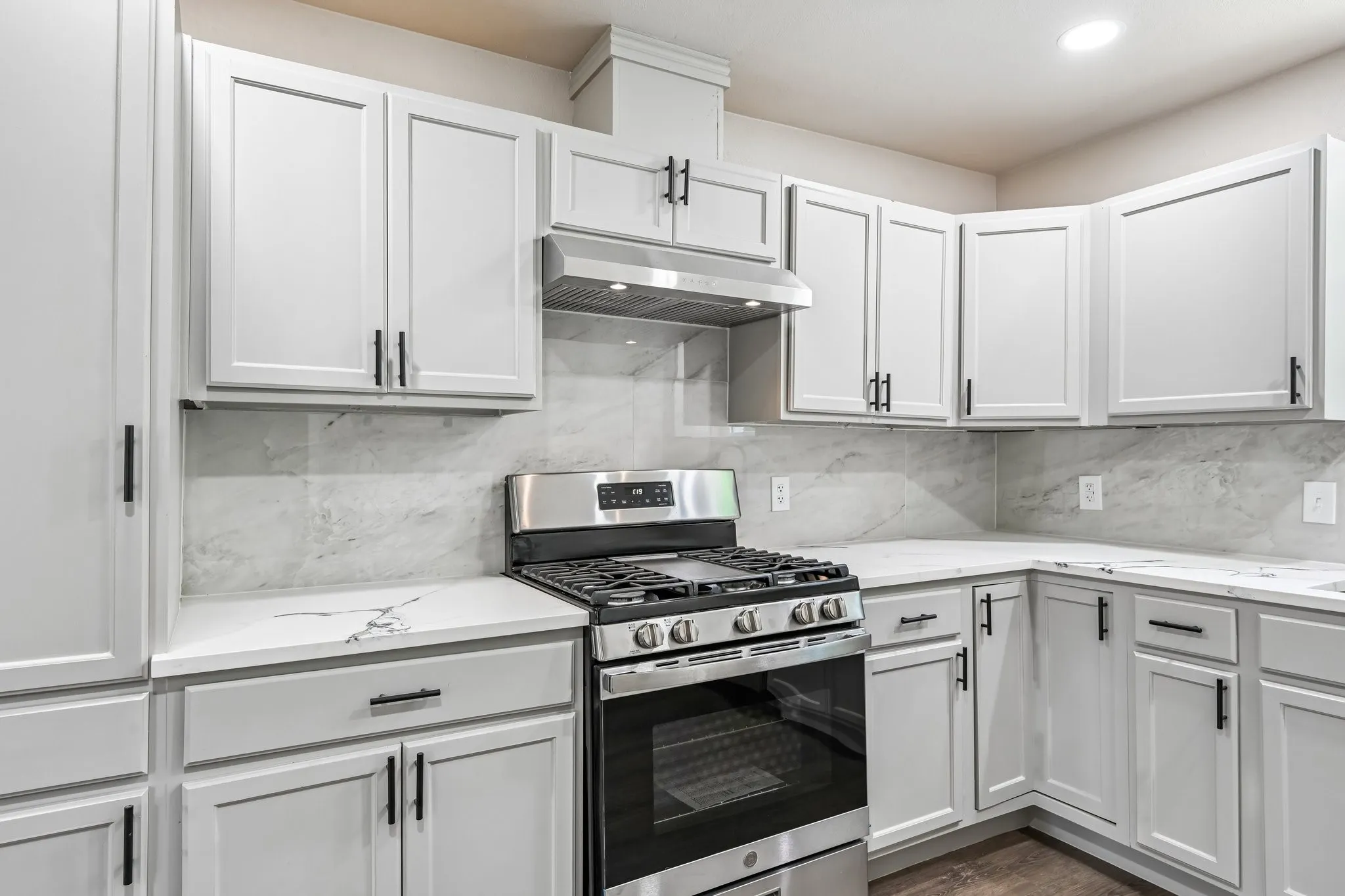 Contemporary kitchen featuring light gray cabinetry with black hardware, white countertops with subtle veining, and a stainless steel gas range with coordinating vent hood
