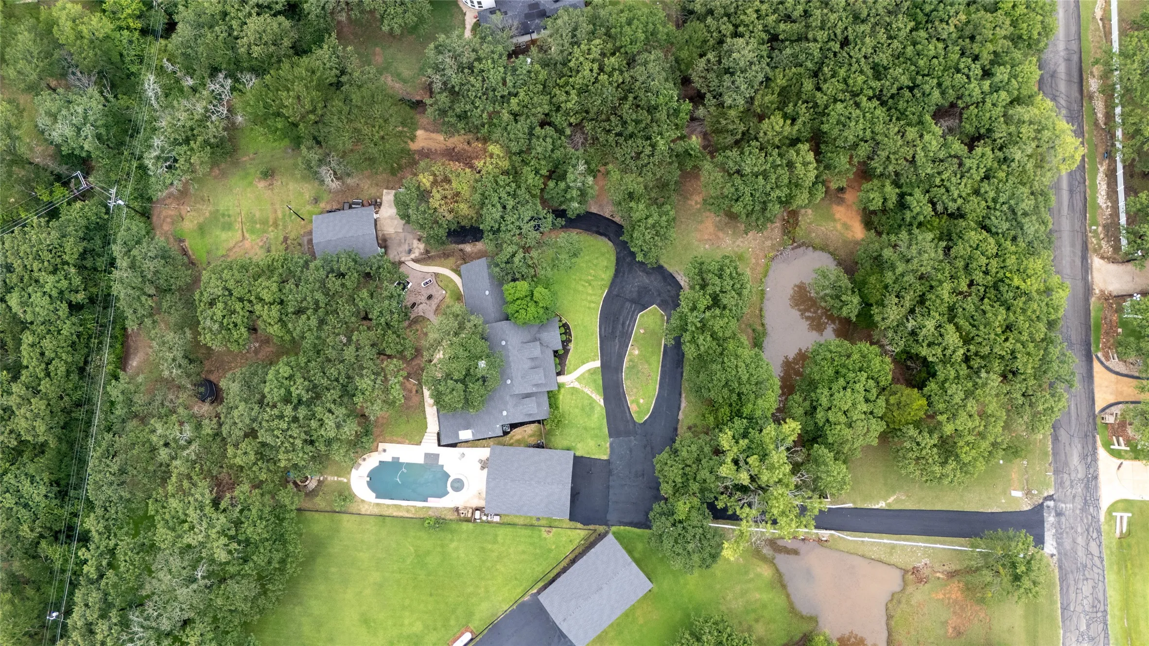 Aerial view showcasing a residential property with established trees, a main residence with a dark-toned roof, a detached structure with a matching roof, a swimming pool with surrounding patio space, and a curved asphalt driveway