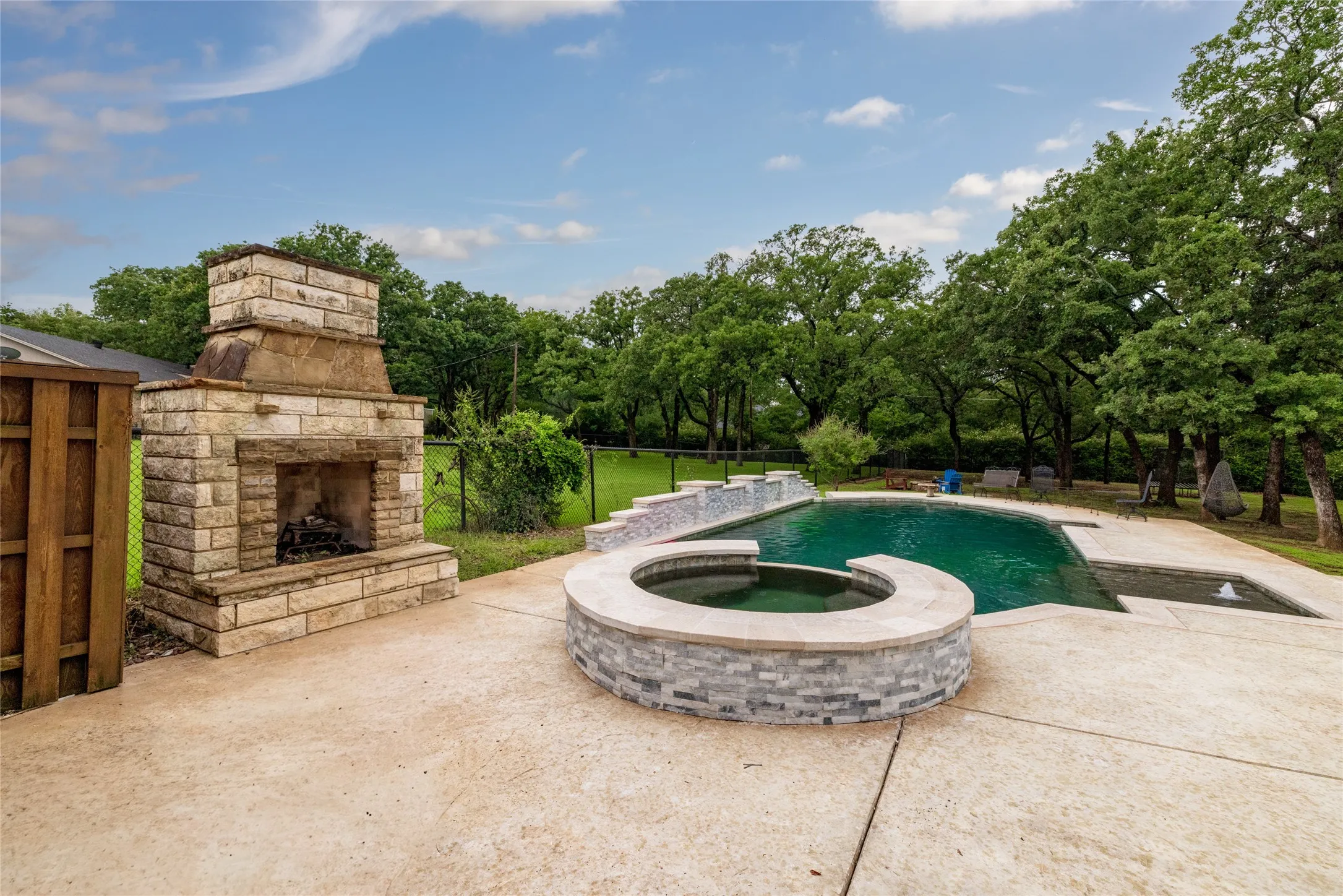 Expansive outdoor patio featuring a built-in stone fireplace, a swimming pool with an integrated spa, and mature trees providing a natural backdrop