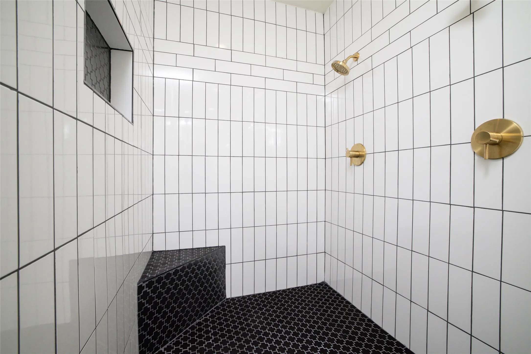Tiled shower featuring vertical white subway tiles, a built-in bench with black patterned tiles, black patterned shower floor tiles, a recessed tile niche, and brass fixtures