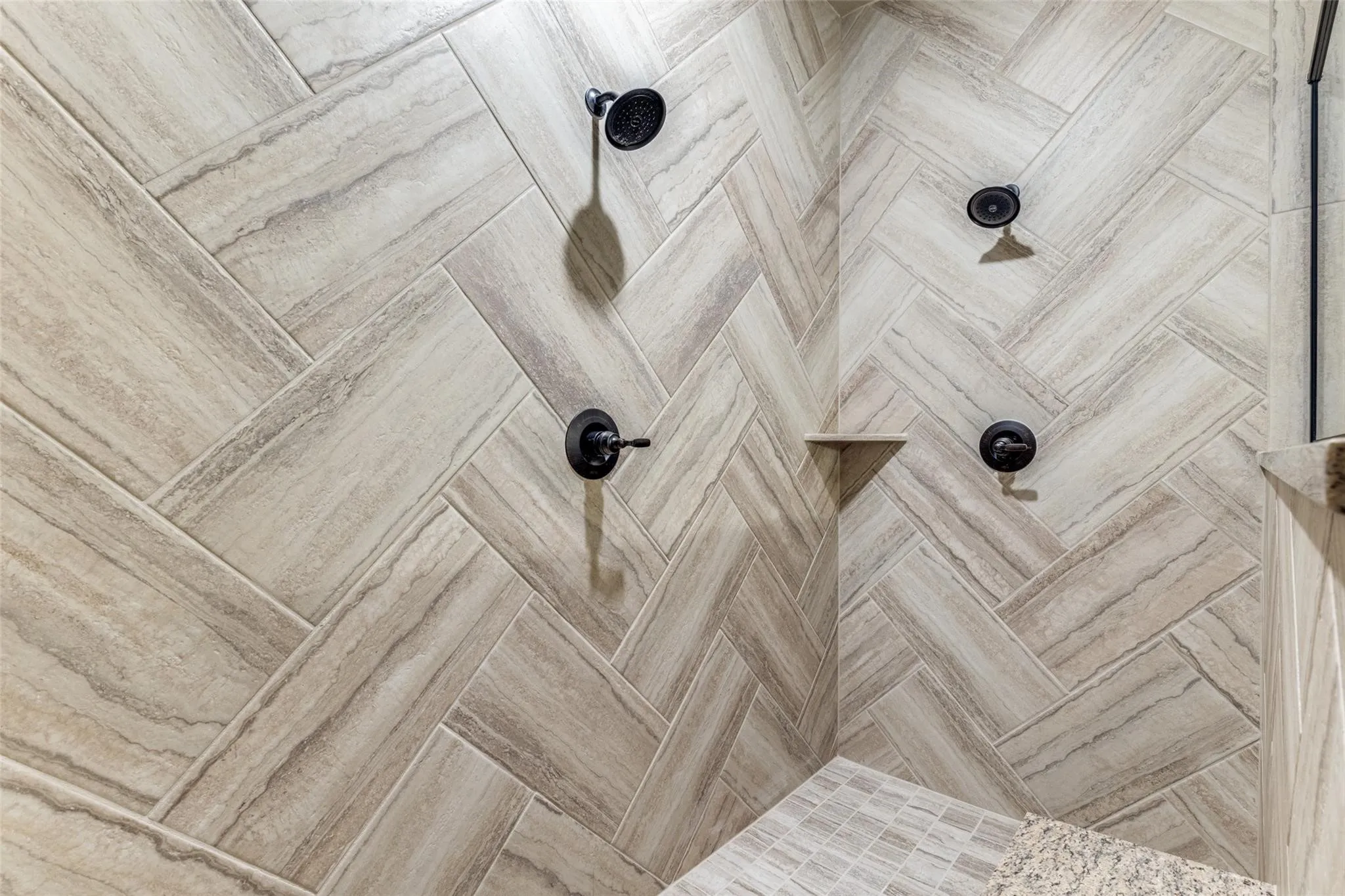 Shower featuring herringbone pattern wall tile, dual matte black shower heads, coordinating matte black controls, an integrated corner shelf, and a tiled shower bench