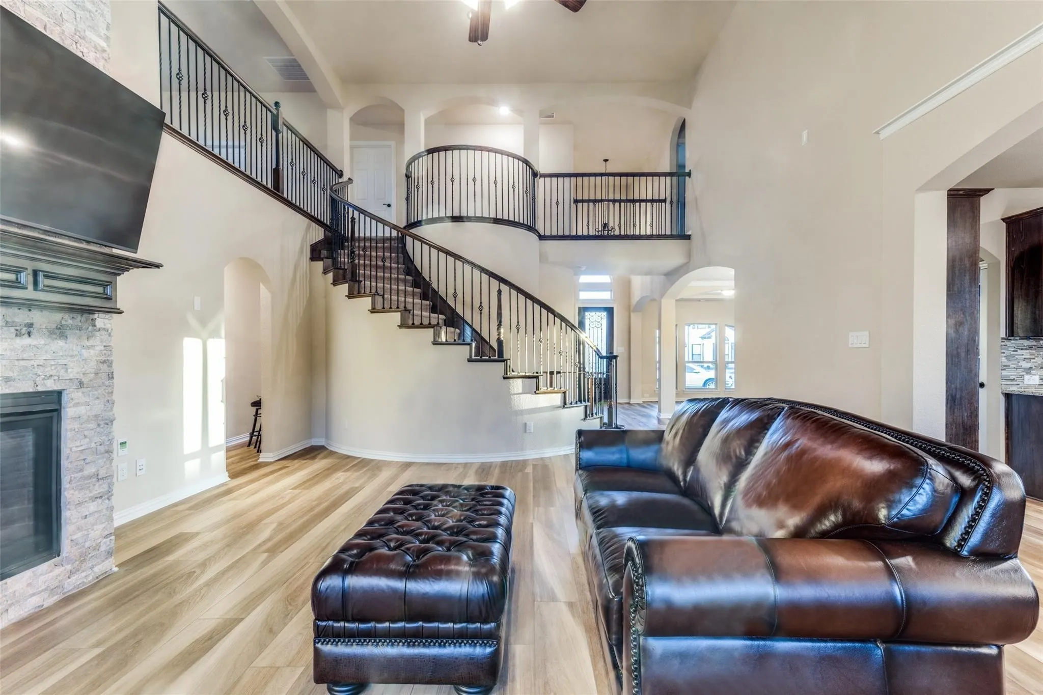 Grand two-story living area featuring a sweeping staircase with wrought iron balusters, a stone-clad fireplace with a decorative mantel, wood-finish flooring, and arched doorways