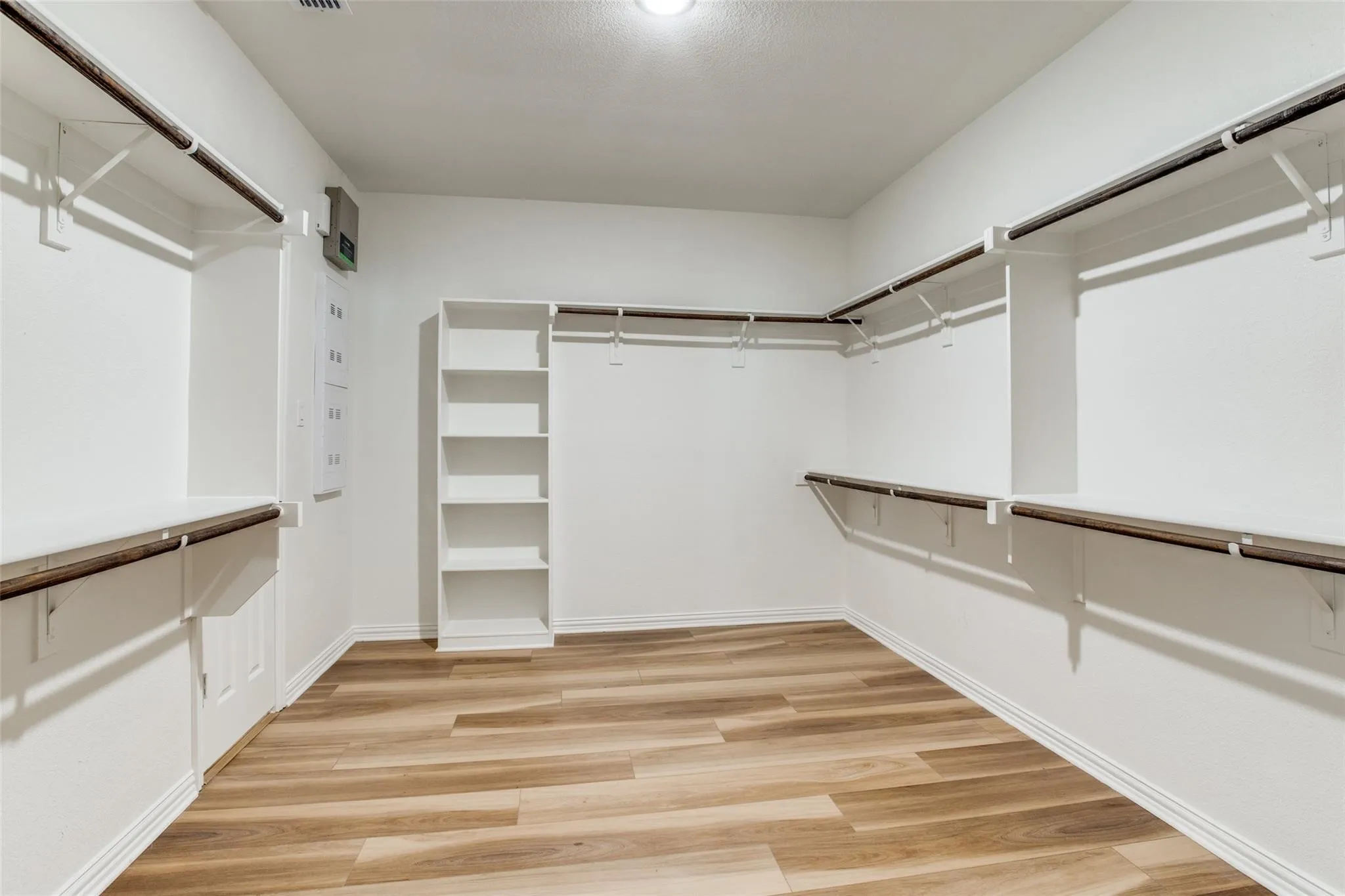 Spacious walk-in closet featuring wood-finish flooring, white walls, built-in shelving, multiple hanging rods, and an overhead light fixture