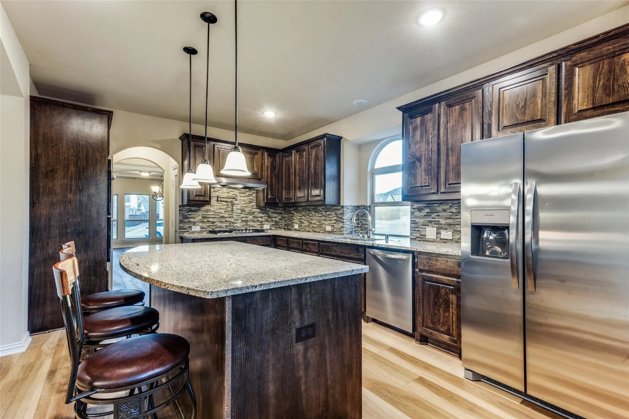 Kitchen featuring a curved island with stone countertop, stainless steel appliances, dark wood cabinetry, a mosaic tile backsplash, and light wood-finish flooring