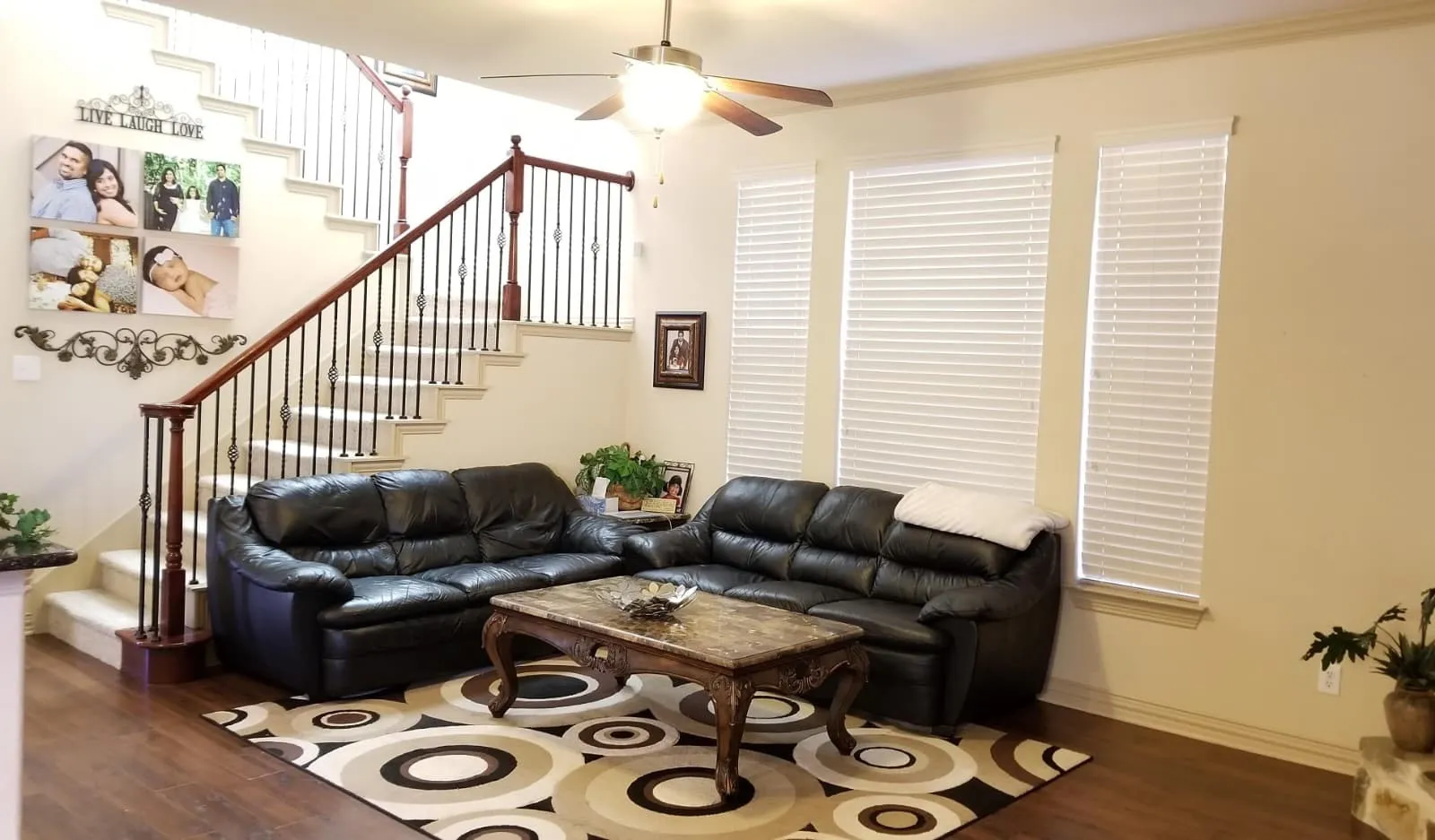 Spacious living area featuring wood-finish flooring, a carpeted staircase with dark wood and wrought iron balusters, and multiple windows with blinds