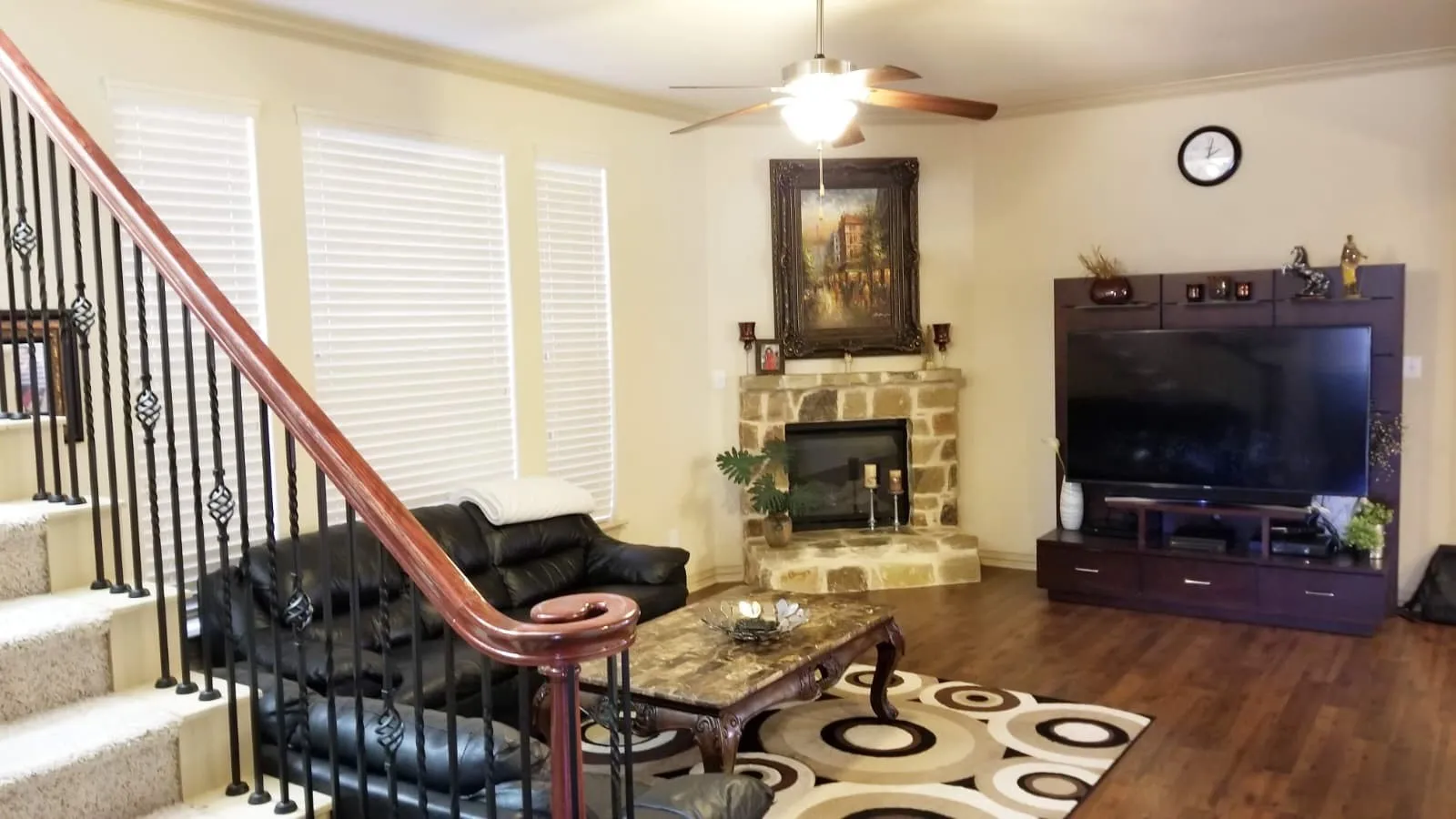 Spacious living area featuring wood-finish flooring, a stone-clad fireplace, and multiple windows with horizontal blinds