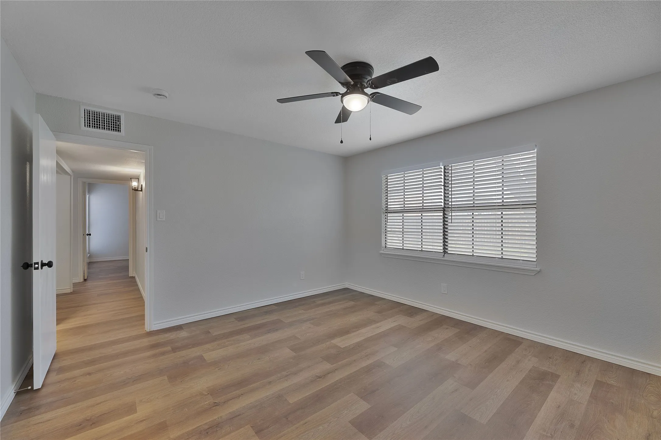 Spacious room featuring wood-finish flooring, a contemporary ceiling fan with integrated lighting, and a window fitted with horizontal blinds
