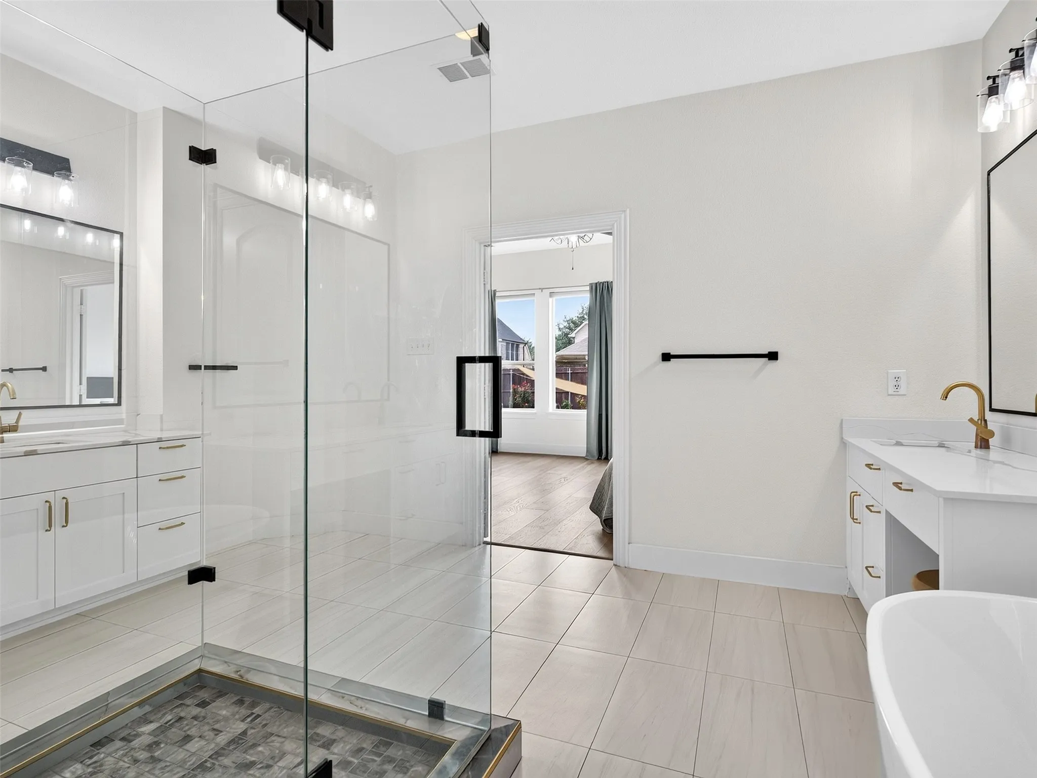 Spacious bathroom featuring a frameless glass shower enclosure, dual vanity sinks with white cabinetry, a freestanding soaking tub, and light-toned tile flooring